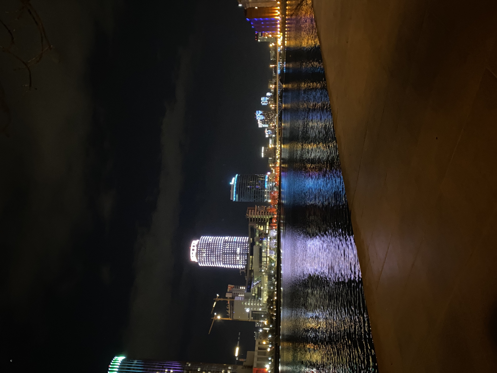 A long exposure photograph showcases a night view of Yekaterinburg, Russia, reflected in a body of water, presumably a river or reservoir. The skyline features multiple lit-up buildings of varying heights, dominated by a tall, illuminated cylindrical skyscraper. The lighting in the city reflects in the water, creating streaks of light. The right side of the image is obscured by an out-of-focus wall or edge, while the left side shows a cloudy sky.