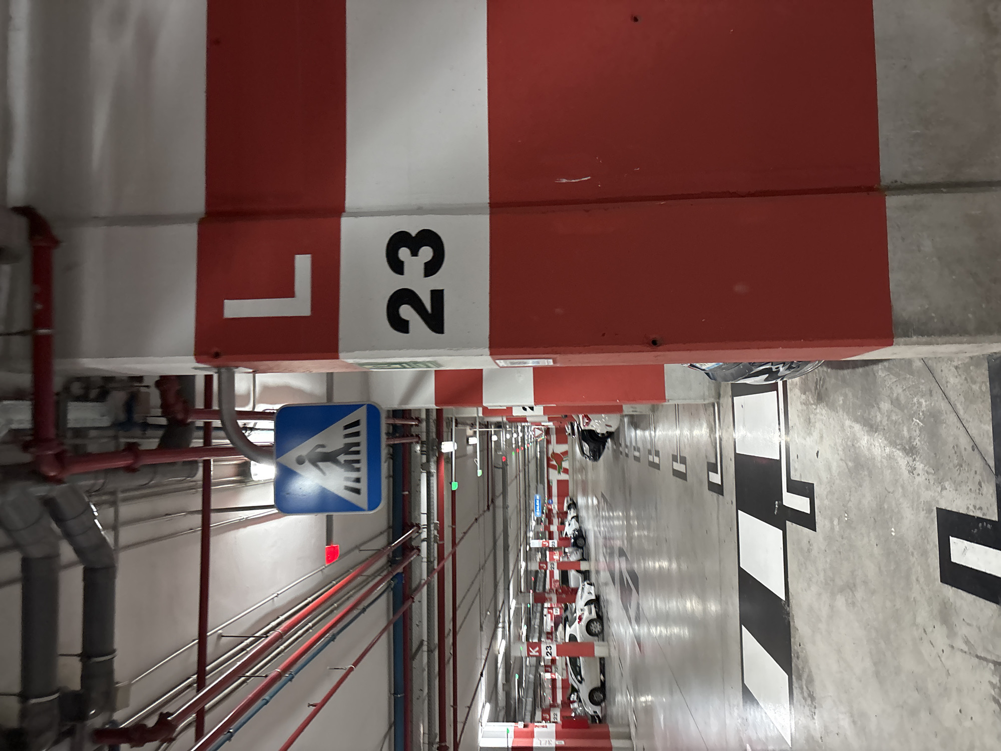 The image depicts a concrete pillar in a parking garage, painted with horizontal red and white stripes. The letter "L" is painted in white on the upper red stripe, and the number "23" is painted in black on the white stripe below. The pillar is located in the city of Orihuela, Spain. In the background, there are cars parked in designated spots, some with red and green lights above them, indicating availability. Overhead, pipes and lighting fixtures are visible, as is a pedestrian crossing sign. The floor is concrete with painted markings for parking spaces.