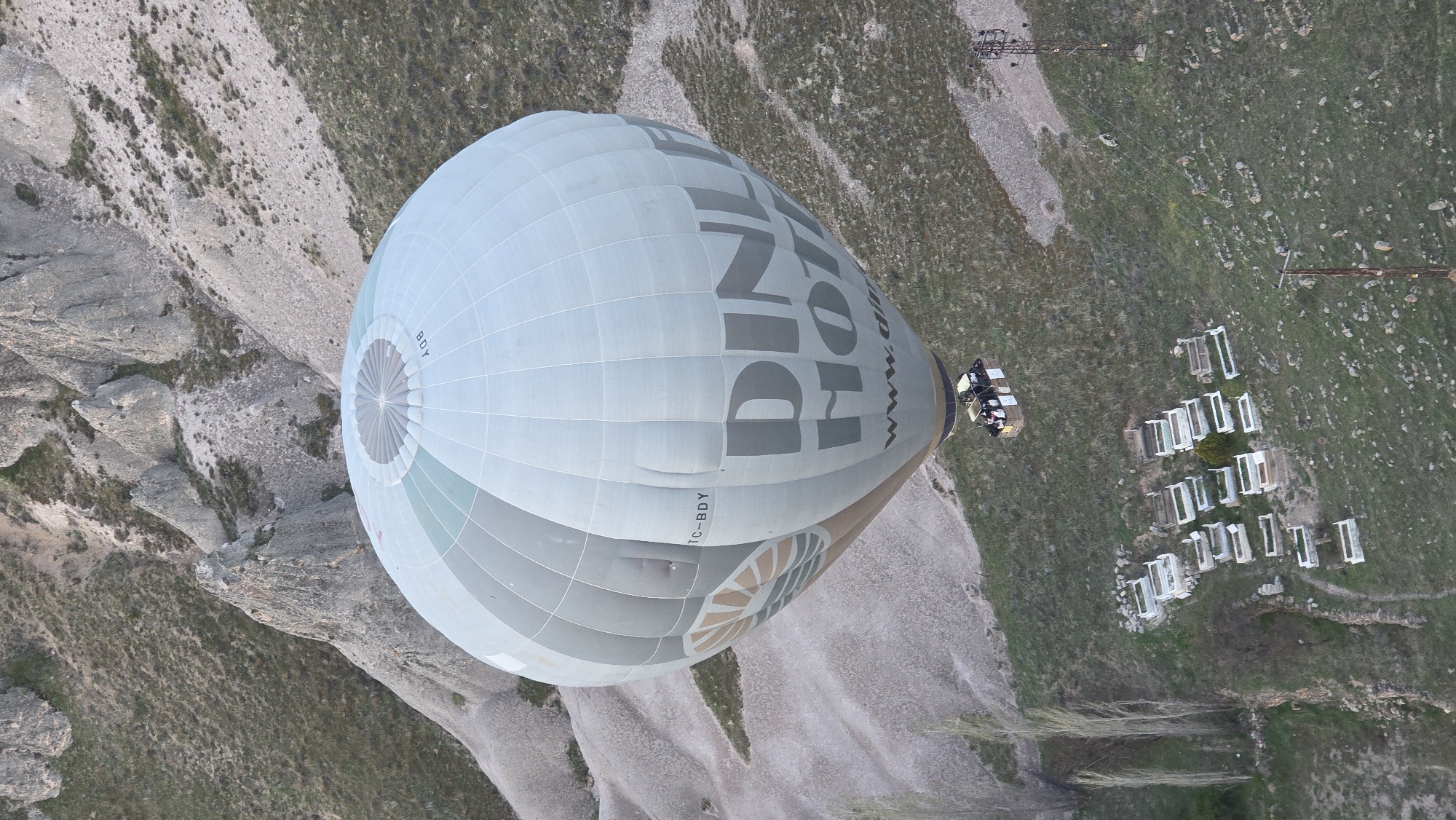 An overhead, slightly tilted shot captures a large hot air balloon in flight over a rugged, arid landscape. The balloon, predominantly light grey and white, features dark grey lettering on its side. Prominently displayed is the word "DİNA", and a partial web address "www.di..." is visible below it. A registration number, "TC-BDY", is also printed on the balloon's surface.
Below the massive balloon, its basket holds a small group of people, participating in the hot air balloon ride. The ground beneath them consists of stark, rocky formations and sparse vegetation, with patches of green contrasting with the lighter-colored soil and rocks. On the right side of the frame, several white rectangular objects, possibly window frames or small shelters, are neatly arranged in rows on a grassy area. Two tall, slender metal structures, resembling poles or towers, are also present in the upper right background, connected by faint lines. The scene is illuminated by soft, natural daylight, indicating clear weather.