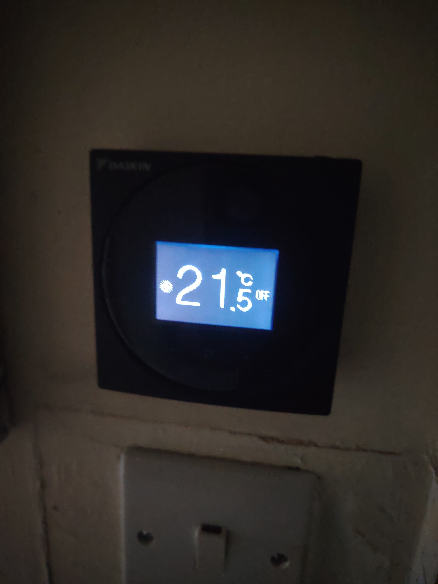 The scene depicts a Daikin brand thermostat mounted on a light-colored wall in a location within Huntingdonshire, United Kingdom. The thermostat is a black, square-shaped device with a central circular element. Its digital display shows "21.5 °C" with a snowflake icon to the left of the numbers, and the word "OFF" in smaller text to the right of the temperature reading. Directly beneath the thermostat, a white, plastic light switch with two visible screws is affixed to the wall, positioned lower in the frame. The wall surface appears to have a slightly textured finish. No people or other active subjects are present in the image.