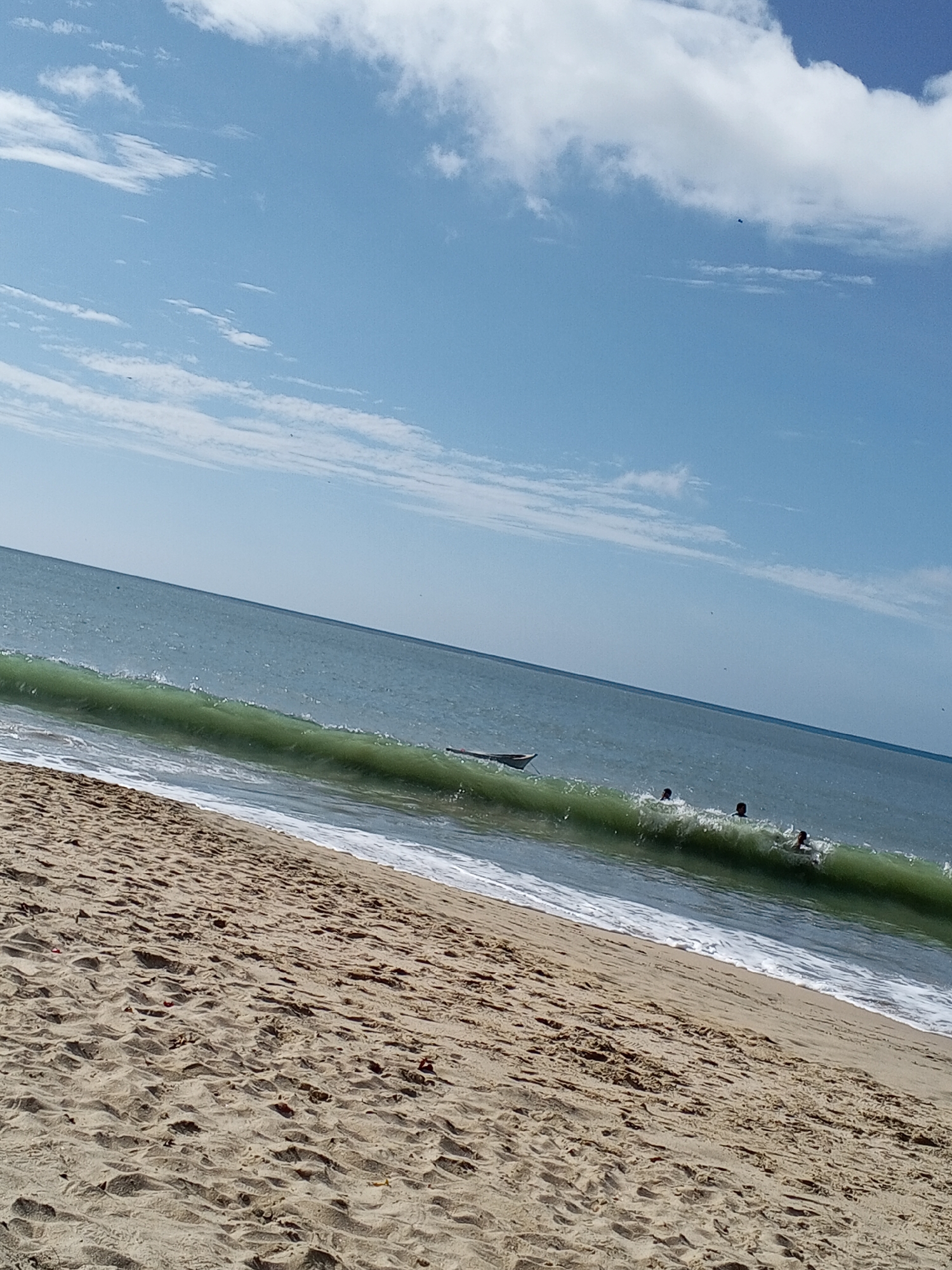 The image depicts a beach scene in Pedro González, Venezuela. A sandy beach occupies the foreground, leading to the ocean. In the water, a wave is breaking, with several people visible within and behind the wave, presumably swimming or surfing. A small boat or vessel is also present near the wave. The sky is blue with scattered white clouds.