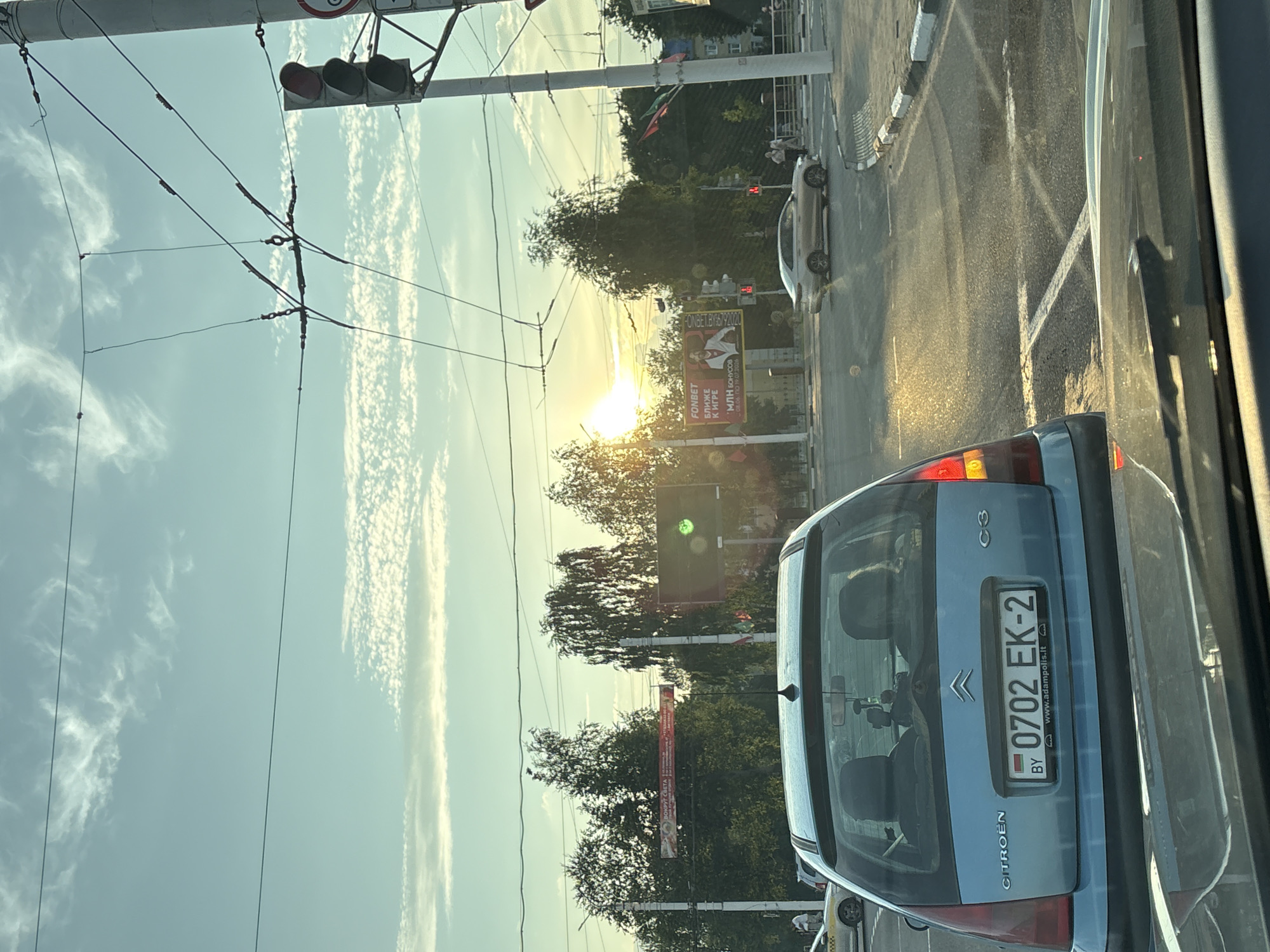 A light blue Citroen C3 with a license plate reading "0702 EK-2" is captured from behind, showing its rear windshield and taillights. The car is stopped at a traffic light intersection under a bright sky with scattered clouds, possibly at sunset. The sun is low in the sky, casting a warm glow on the road and surrounding trees. In the background, a billboard advertising "FONBET" is visible, along with traffic lights and overhead power lines. Other vehicles and pedestrians are present in the distance, indicating an urban environment. The license plate's format and the flag emblem suggest the car is registered in Belarus, specifically from the region of Vitsebsk.