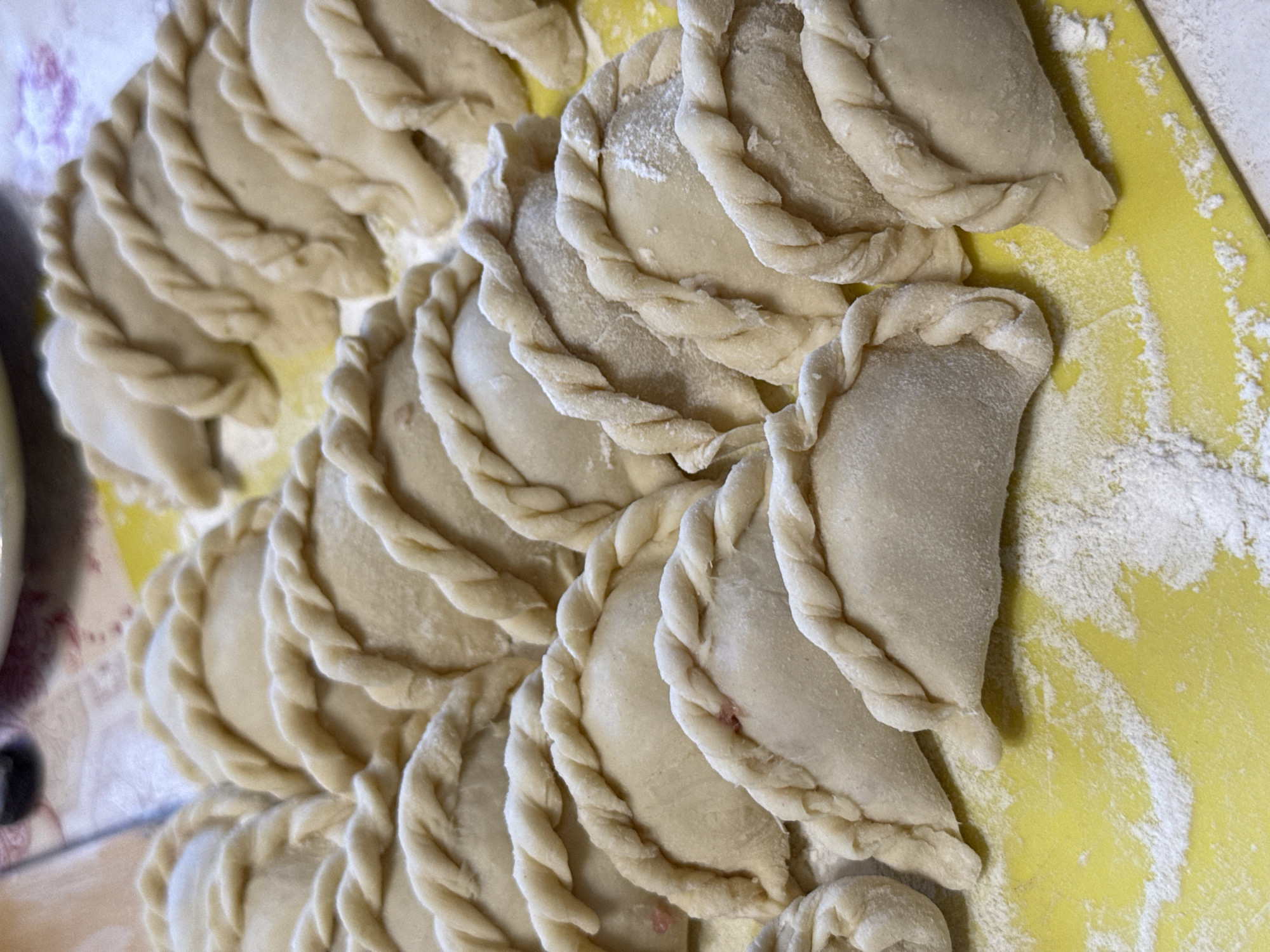 The image shows a close-up of numerous uncooked pierogi, arranged on a yellow surface. The pierogi are crescent-shaped, made of dough, and have crimped edges. Some flour is scattered on the yellow surface. The pierogi are not cooked. The location is not confirmed.