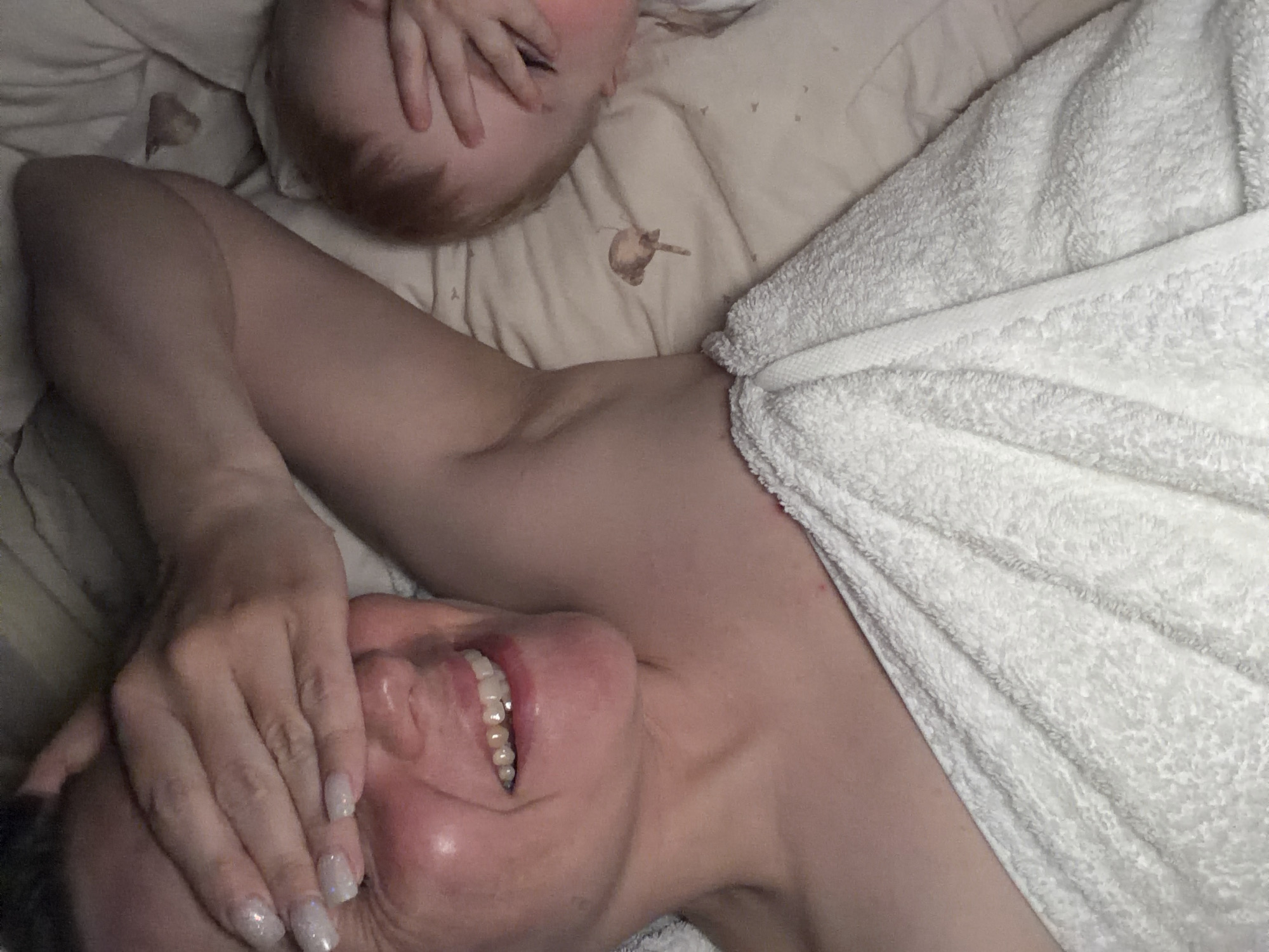 A woman lies on her back in a bed, partly covered by a white towel. Her face is visible, with her hand covering her forehead. A baby lies next to her on the bed, their face obscured by their hand. The bed has light-colored sheets. A small, unidentified object rests on the sheet near the baby. The location is unknown and cannot be determined from the image.