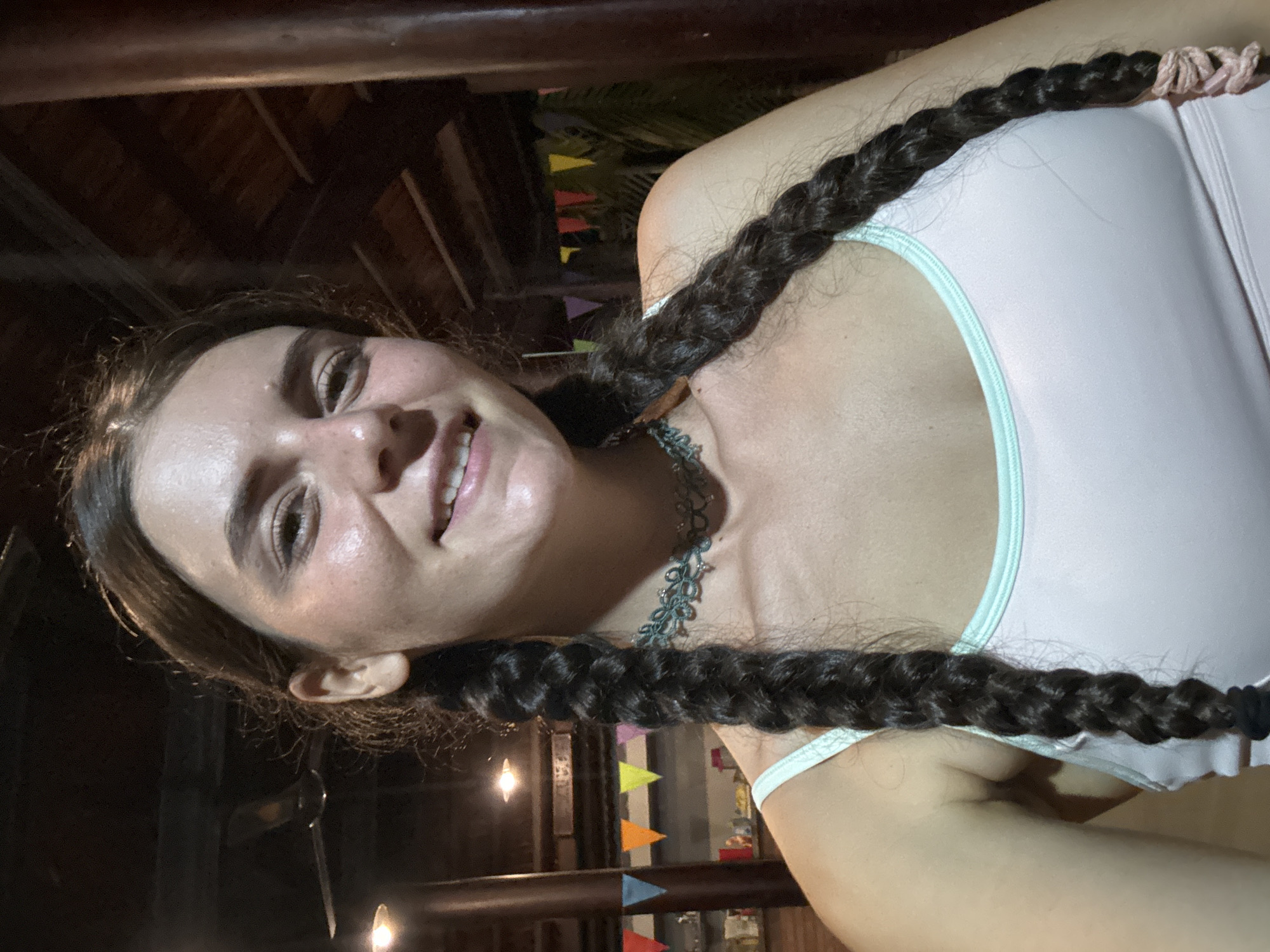 A selfie of a young woman with dark hair in two long braids, taken in Mũi Né, Vietnam. She is wearing a light pink tank top with light green trim and a dark green or grey necklace. The woman is smiling and looking directly at the camera. The background is blurred but shows some colorful flags, lights, and wooden structures. It appears to be an outdoor setting, possibly at a restaurant or bar.