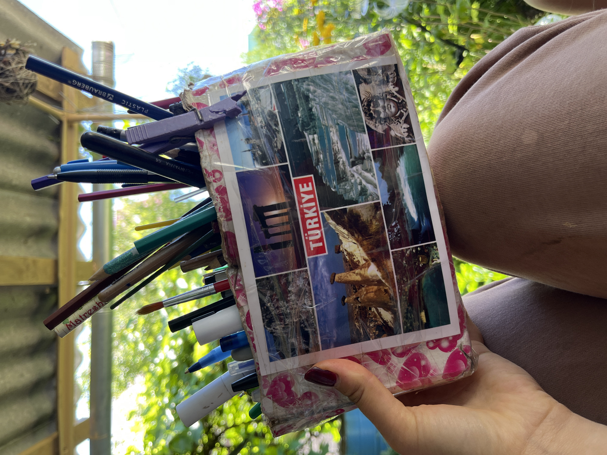 A person's hand, with dark red nail polish, holds a handmade pen and pencil holder. The holder is a rectangular box, possibly made from repurposed materials, covered with clear tape and decorated with a collage of images of Turkish landscapes and landmarks, including the word "TÜRKİYE" in red lettering. The holder is filled with various writing and drawing instruments, such as pens, pencils, and brushes, some of which are visible at the top. The background suggests an outdoor setting with lush greenery and sunlight filtering through a roofed structure, possibly a veranda or gazebo. The scene is likely located in a private outdoor space, not a public area in the city of Dunilavichy, Belarus, as there are no identifiable urban elements.