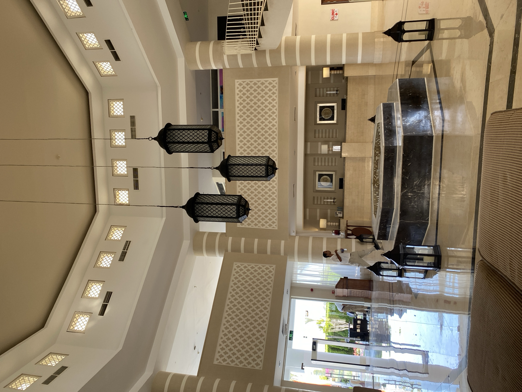 This image captures the interior of a spacious, modern hotel lobby in Sharm El Sheikh, Egypt, characterized by its bright, airy ambiance and elegant design. The lobby features a high, multi-tiered ceiling with recessed lighting and decorative square panels. Dominating the space are three large, dark-colored lanterns hanging from the ceiling by thin cords, adding a touch of ornate flair.
The floor is made of highly polished marble tiles in light beige and darker brown, reflecting the ambient light and the architectural elements. In the center of the lobby is a hexagonal water feature with dark marble cladding, surrounded by patterned floor tiles. Two additional black lanterns stand on the floor near the water feature.
The walls are a neutral beige color, adorned with geometric patterned panels. Large columns, also beige with white rings, support the ceiling structure. To the left, a large entrance leads to an outdoor area with greenery visible. A man in a light-colored shirt and pants is seen walking away from the camera in the mid-ground, holding a bottle of water. To the right, a staircase with a white railing curves upwards, leading to an upper level. Further to the right, a sign indicates a "CLINIC".
The overall impression is one of a well-maintained and luxurious hotel environment, typical of resorts in Sharm El Sheikh, designed to impress guests upon arrival.