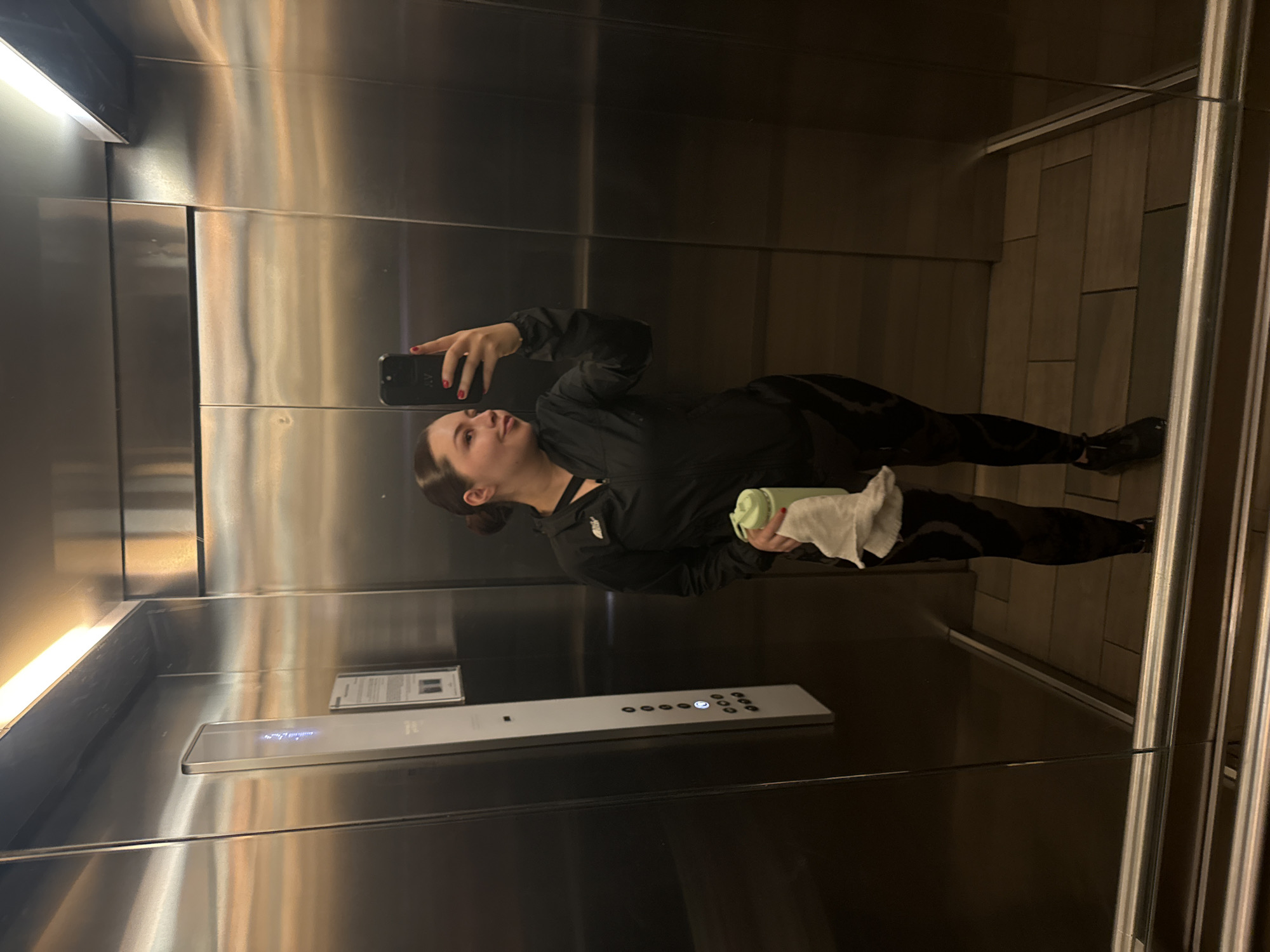 A young woman is captured taking a selfie in an elevator, holding her smartphone up with her right hand. Her dark hair is pulled back, and she wears a black athletic jacket with a white logo partially visible on the front, along with dark, patterned leggings and dark shoes. Her fingernails are painted red. In her left hand, she holds a light green water bottle and a folded white towel. She is looking upwards and slightly to her right, towards the phone screen.
The elevator interior features highly polished, reflective metallic walls that show distortions of the woman's reflection and the ambient light. The floor is covered with rectangular, light brown wooden panels arranged in a staggered pattern. A long, narrow light fixture runs along the top of one wall, casting a soft glow. A sleek, silver-colored control panel is visible on the wall opposite the woman, featuring multiple circular buttons. One button is illuminated, and the letters "LG" can be faintly seen on another button. Above the control panel, a small rectangular sign with unreadable text is mounted. The overall lighting suggests an indoor setting. The specific location is Mexico City, Mexico.