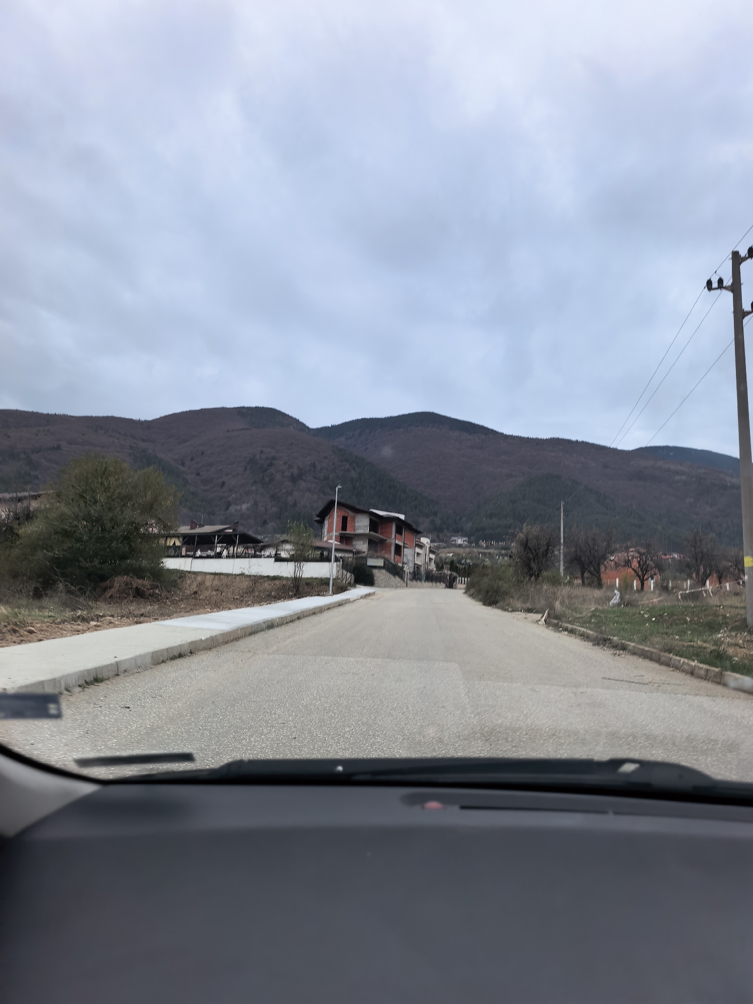The image shows a view from inside a car, looking out onto a paved road that leads uphill. There are mountains in the background under a cloudy sky. A partially constructed brick building is on the left side of the road, and there is a utility pole on the right side. The location appears to be a residential area, possibly on the outskirts of a town.