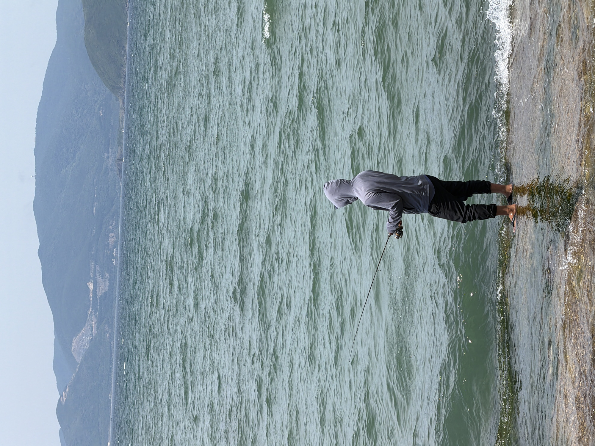 A person stands in shallow water at the edge of a body of water, fishing. They are wearing a grey hooded sweatshirt and dark pants. The water is rippling and there are waves breaking near the shore. In the background, there are mountains. The location is identified as Bac Nha Trang Ward, Vietnam.