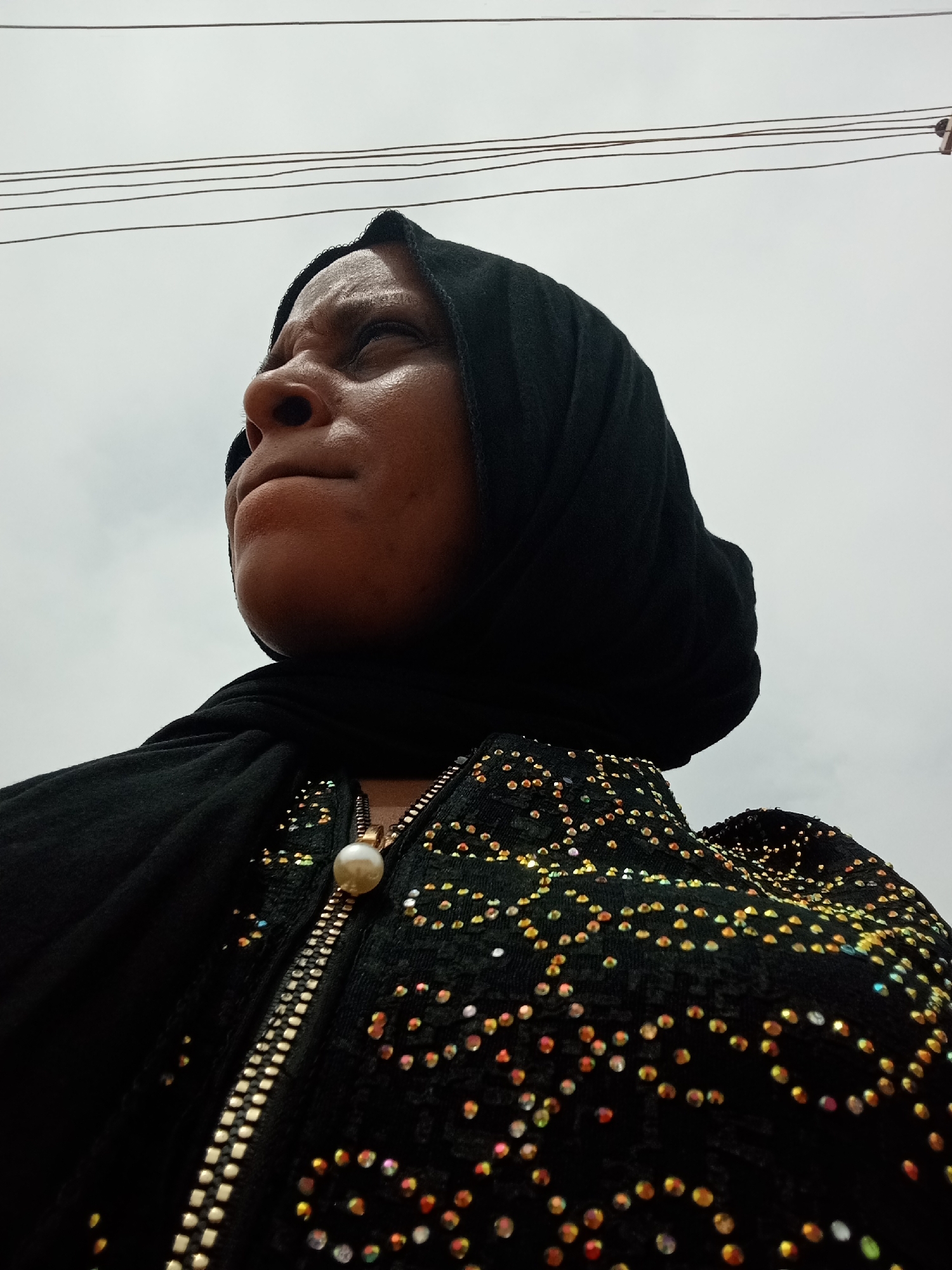 A woman with dark skin wears a black headscarf and a black sequined top with a zipper. She is looking off to the left side of the frame. Overhead are electrical wires against a pale sky. The location is Narayi, Nigeria.