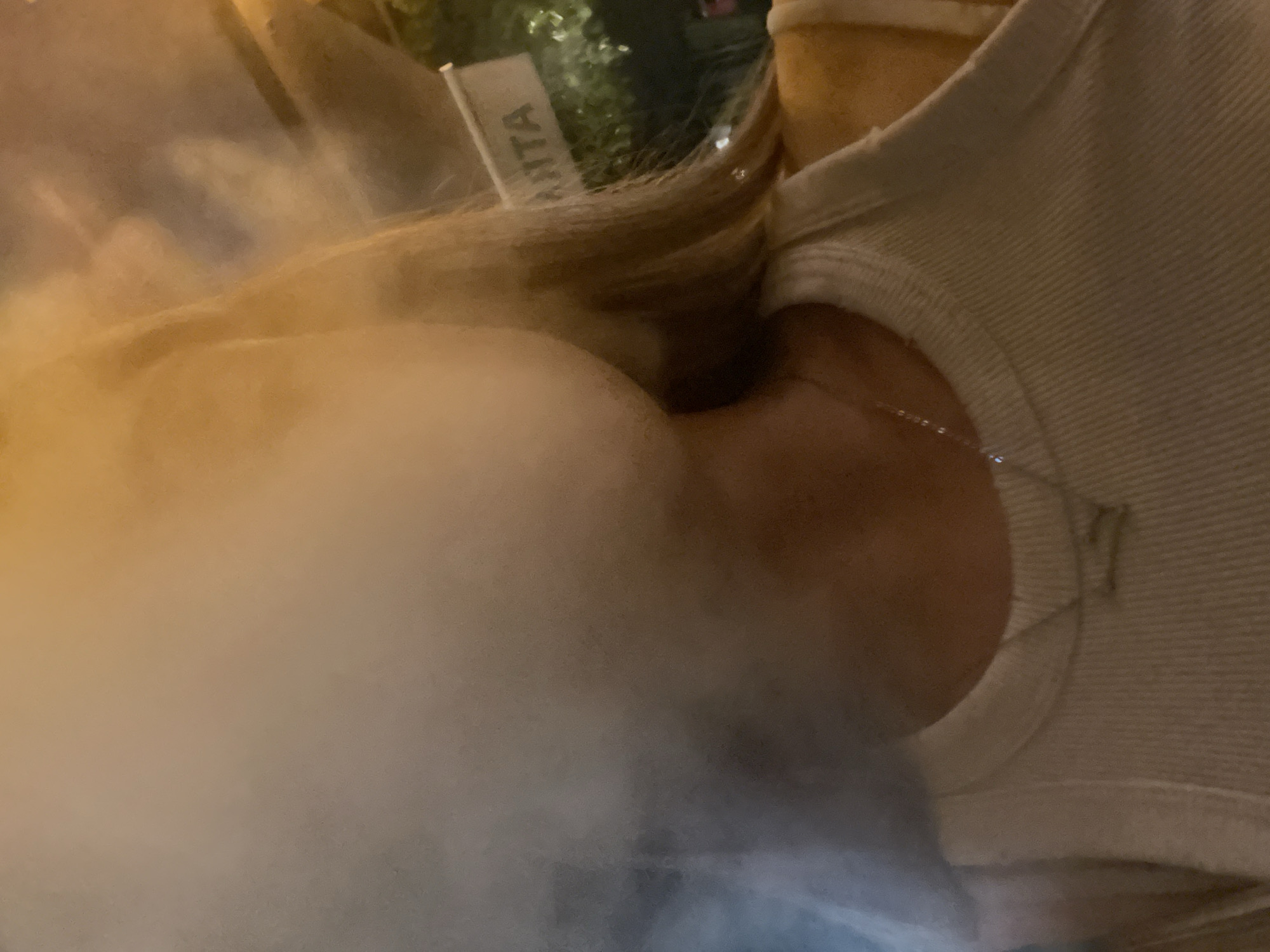 A forensic close-up captures a person exhaling a large volume of white vapor, obscuring their face, against a dark, ambiguously lit background in the city of Unknown, Unknown. The subject, identifiable by blonde hair partially visible on the right side, wears a light-colored ribbed tank top and a thin silver necklace with a small pendant. The dense vapor occupies the left and central portions of the frame. In the upper right background, a white sign with dark lettering, partially displaying "ANTA", is visible, with dark foliage and blurry lights behind it, suggesting an outdoor nocturnal setting. The lighting is primarily warm, originating from the right side of the frame, illuminating the vapor.