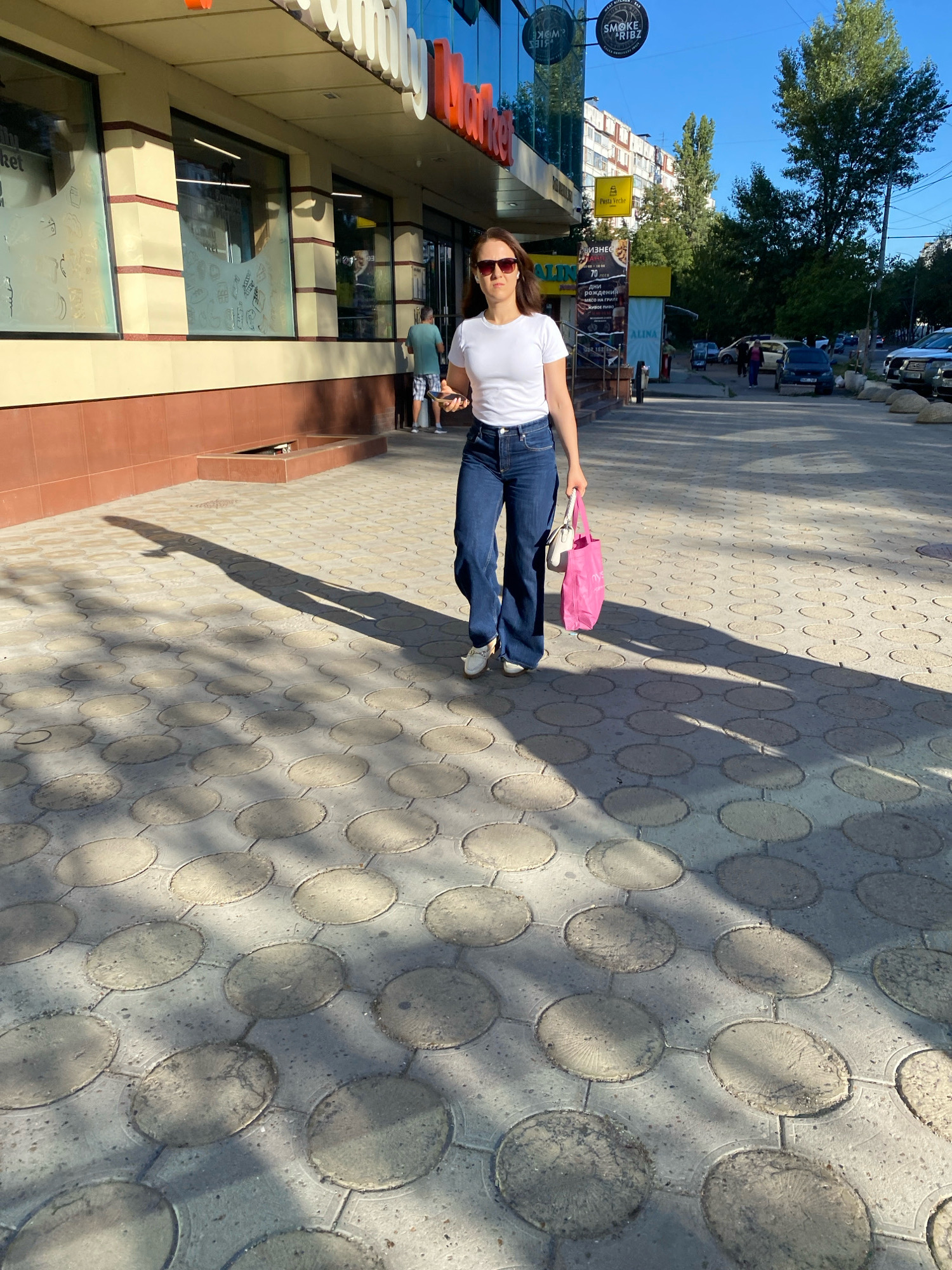 A woman walks on a sidewalk in front of a storefront. She is wearing a white t-shirt, blue jeans, sunglasses, and white shoes. She is carrying a pink tote bag and a white purse. The storefront has large windows and a red brick base. The sidewalk is paved with hexagonal concrete tiles. The sun is shining, casting shadows on the sidewalk. In the background, there are cars, trees, and buildings. The image appears to be taken in Chișinău, Moldova, as suggested by the Cyrillic lettering on the storefront, which reads "Family Market".
