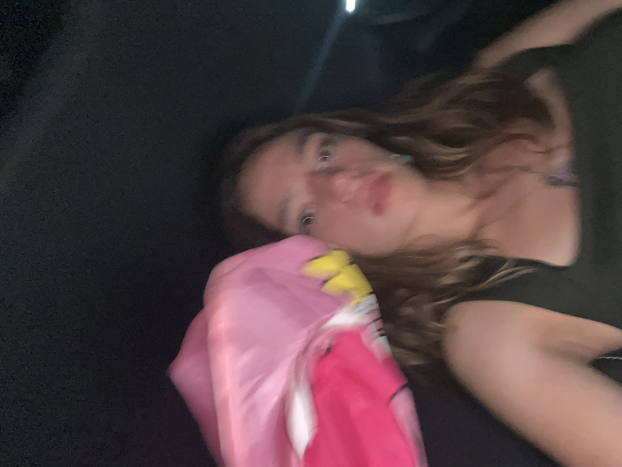 A woman with light skin and long, wavy brown hair is lying down, possibly inside a vehicle due to the dark background and glimpses of the vehicle's interior. She is wearing a dark-colored shirt and has a pink and yellow item positioned near her head, which could be a pillow or a piece of clothing. The image is somewhat blurred. The location is unknown.