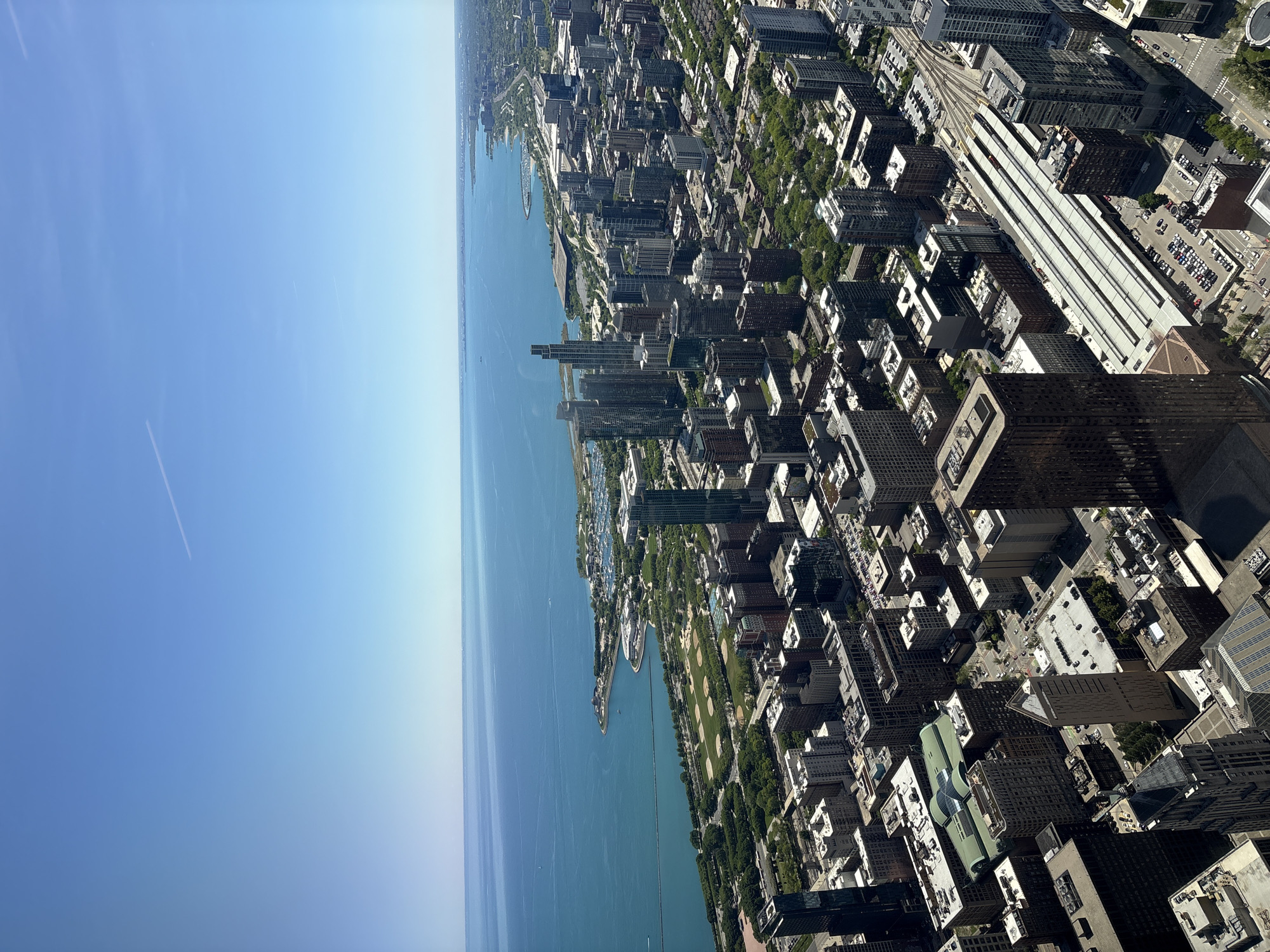 The image depicts an aerial view of Chicago, Illinois, USA, on a clear day. Lake Michigan is visible on the left, with a blue sky above. The city skyline is composed of various buildings, including skyscrapers and residential structures, concentrated along the shoreline. There is visible green space, probably park area, as well as roads and railways. The buildings appear densely packed, indicating a typical urban environment.