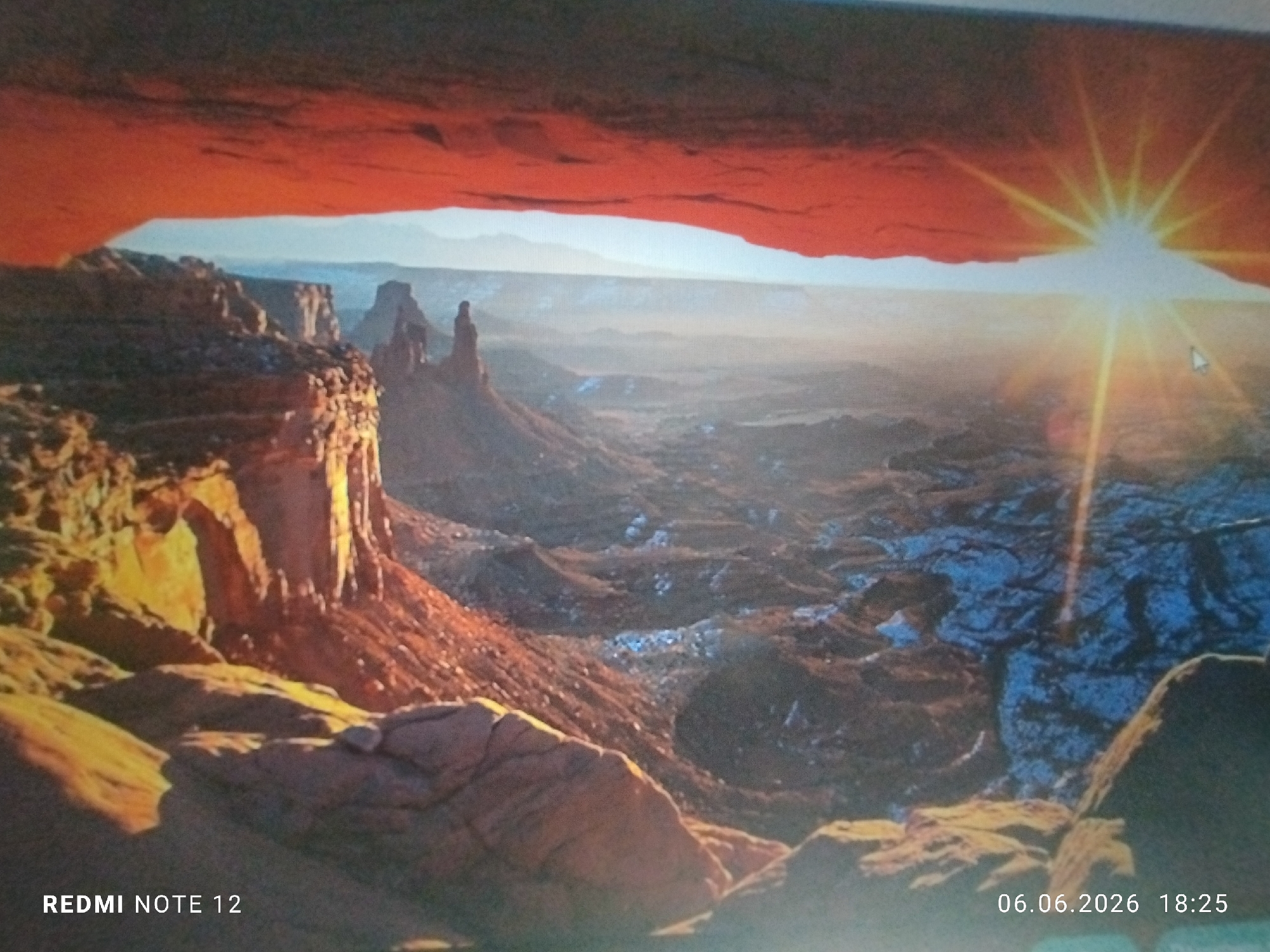 The image is a photograph taken in the city of Burgas, Bulgaria, depicting a digital screen displaying a sunrise landscape over a vast canyon. The scene on the screen centers on a prominent reddish-orange sandstone arch, identified as Mesa Arch, which frames a panoramic view of the canyon below. Through the arch, a deep canyon stretches into the distance, characterized by numerous eroded rock formations, including distinct mesas and pinnacles, all illuminated by the warm light of a rising sun. The sun is positioned above the horizon on the right side of the displayed image, emitting strong, star-like rays and a bright glare. The canyon floor and surrounding slopes are predominantly reddish-brown, with visible patches of white, likely snow or frost, concentrated in shaded areas, particularly in the mid-ground towards the right. The immediate foreground of the screen's display shows additional rugged, multi-toned rock formations. A white computer mouse cursor is present on the screen, positioned in the middle-right quadrant, pointing towards the sun. In the bottom-left corner of the photograph, the text "REDMI NOTE 12" is visible, and in the bottom-right corner, the timestamp "06.06.2026 18:25" is displayed, indicating the camera model and the date/time of the photograph taken of the screen. No human subjects or discernible actions are depicted within the landscape itself.