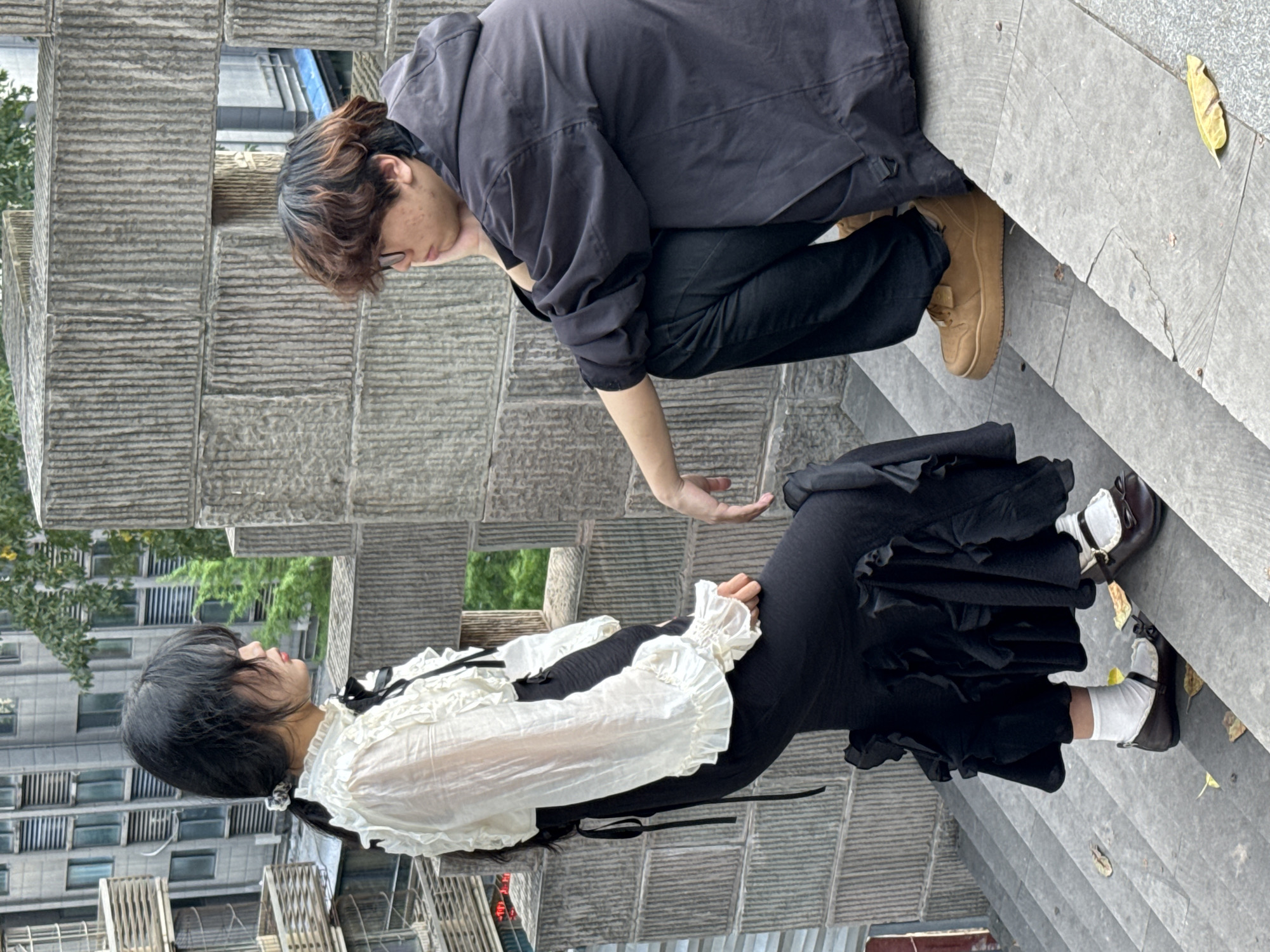 Two people are interacting on some steps in the Yuzhong District, China. One person, wearing a dark jacket, black pants, and tan shoes, is bending over towards another person. The second person is wearing a white frilly top, a black skirt with ruffles, white socks, and dark shoes with straps. They are positioned on a set of stairs with fallen leaves. The background includes a building with a textured facade and glimpses of green trees. A nearby structure with a blocky design is present.