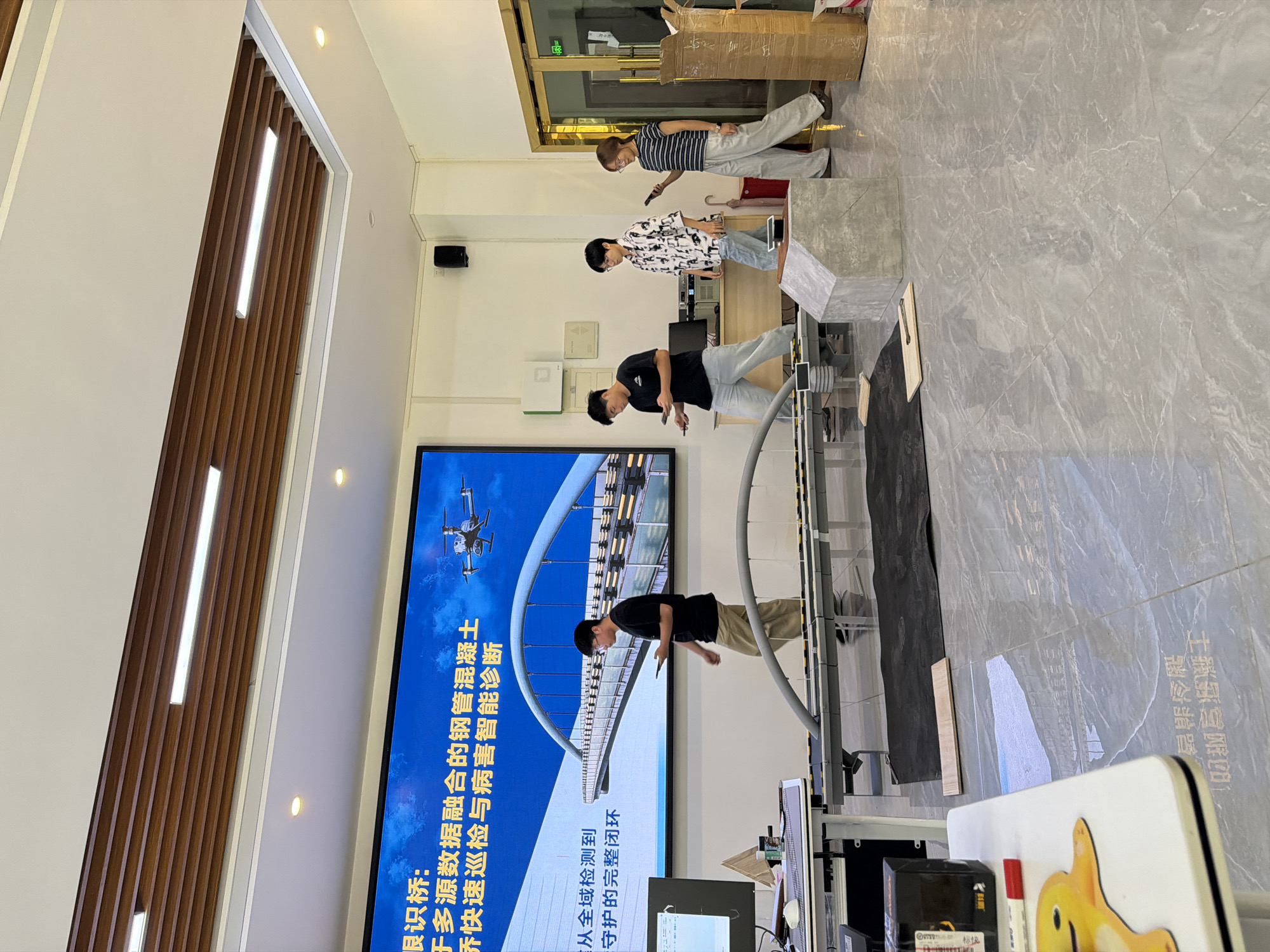 The image depicts an indoor scene, possibly a presentation or demonstration area. A large digital screen displays text and an image of a bridge with a drone, suggesting a technical or engineering context. Four individuals, appearing to be young adults, are present in the room. One person on the left is looking at a laptop. Three other individuals are walking and looking at their mobile phones. In the foreground, a model of a bridge structure is set up on a table, and there are various objects like boxes, a concrete block, and a red marker on the floor and tables. The environment features a modern interior design with a tiled floor and wooden ceiling panels. There is no visual information in the image that allows for the identification of the city as Jiangjin, China, or any specific urban location.