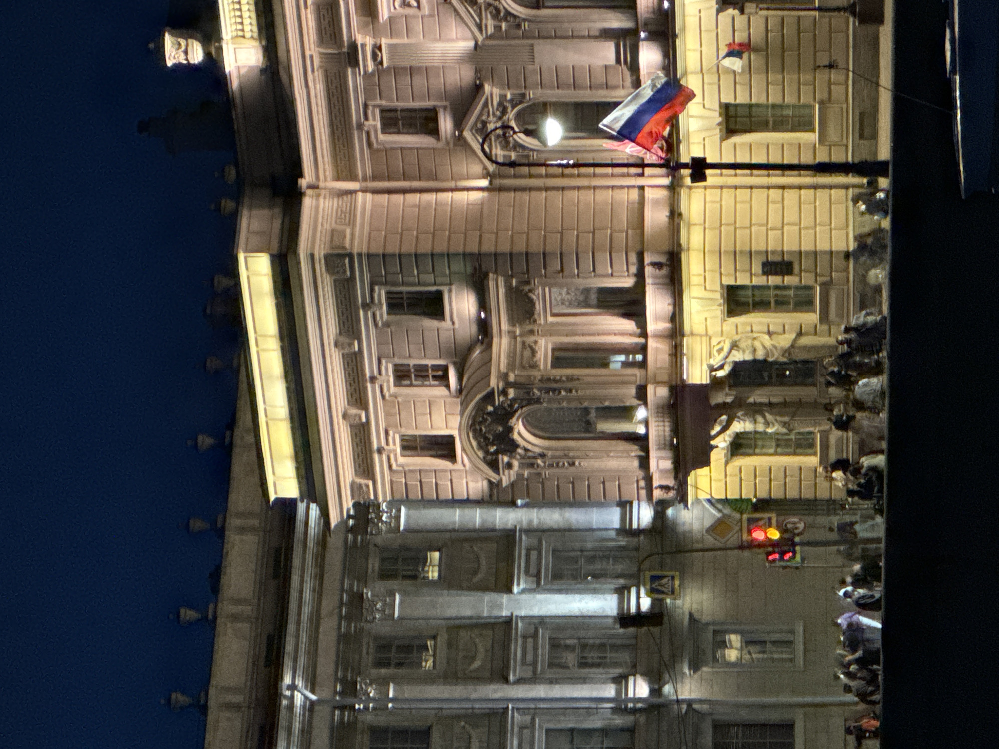 The image depicts a nighttime scene of the Palace Bridge in Saint Petersburg, Russia, illuminated by artificial lights. The architectural style of the buildings suggests a classical or neoclassical design, characterized by columns, ornate windows, and decorative elements. A Russian flag is prominently displayed on a flagpole, adding a nationalistic element to the scene. Several people are visible in the foreground, appearing to be tourists or residents enjoying the evening. Some are gathered in groups, while others are walking or standing individually. The presence of streetlights and traffic signals indicates an urban environment. The dark sky and the illuminated buildings create a dramatic contrast, highlighting the grandeur of the architecture. The overall impression is one of a lively and picturesque urban setting at night.