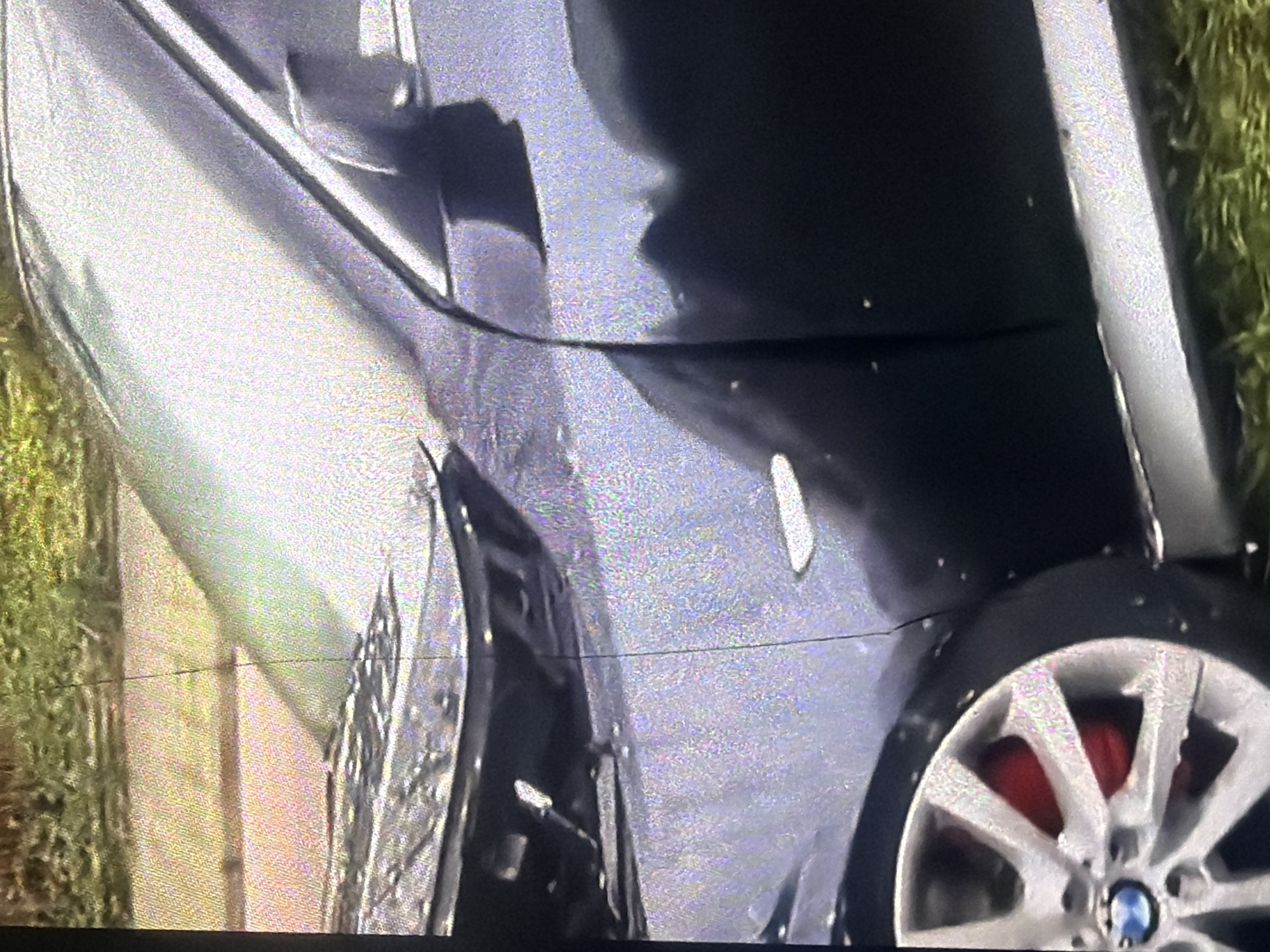This image captures a damaged dark-colored sedan, likely a BMW based on the wheel emblem, parked on a street in Bukuru, Nigeria. The front of the car is heavily damaged, with the hood appearing crumpled and misaligned. The driver's side door and front fender show signs of impact, with visible scuffs and potential deformation. A portion of the car's tire and a red brake caliper are visible in the foreground. The surrounding environment appears to be a grassy area next to a paved surface, possibly a sidewalk or road shoulder. No individuals are present in the image. The lighting suggests it is daytime.