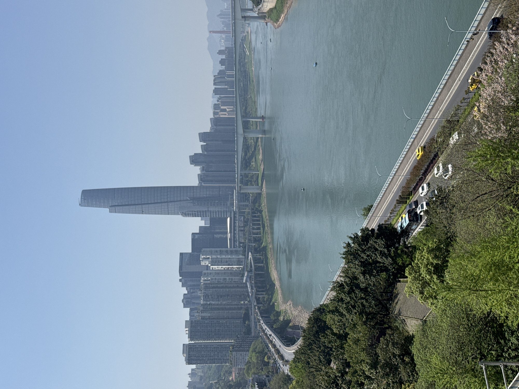 The image depicts a daytime aerial view of the Yuzhong District in Chongqing, China. A large river runs diagonally through the frame, with a bridge spanning it. A modern skyline of tall buildings rises along the riverbank. A prominent, uniquely shaped skyscraper stands out. Trees line the river bank. A road runs adjacent to the river with vehicular traffic, including cars and buses. The sky is clear.