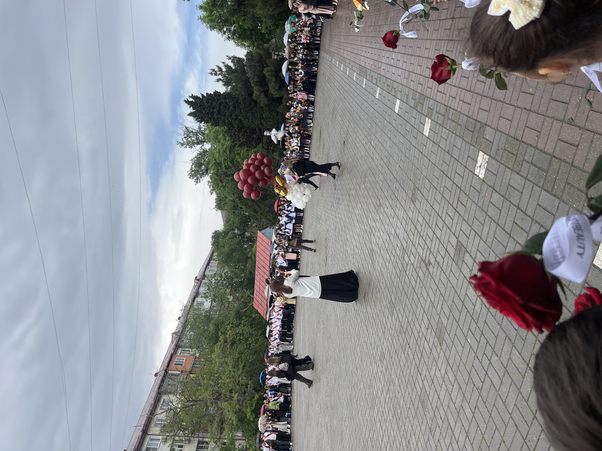 The image shows an outdoor event in the city of Kaspiysk, Russia, likely a graduation ceremony. A large group of people are lined up along a brick-paved area, presumably students. A woman in a white top and black skirt is facing the crowd and holding her hand up. Two individuals run towards the center of the brick area carrying balloons. Along the ground are roses. Buildings and trees form the backdrop, and power lines stretch across the sky. The sky is overcast, suggesting a cloudy day.