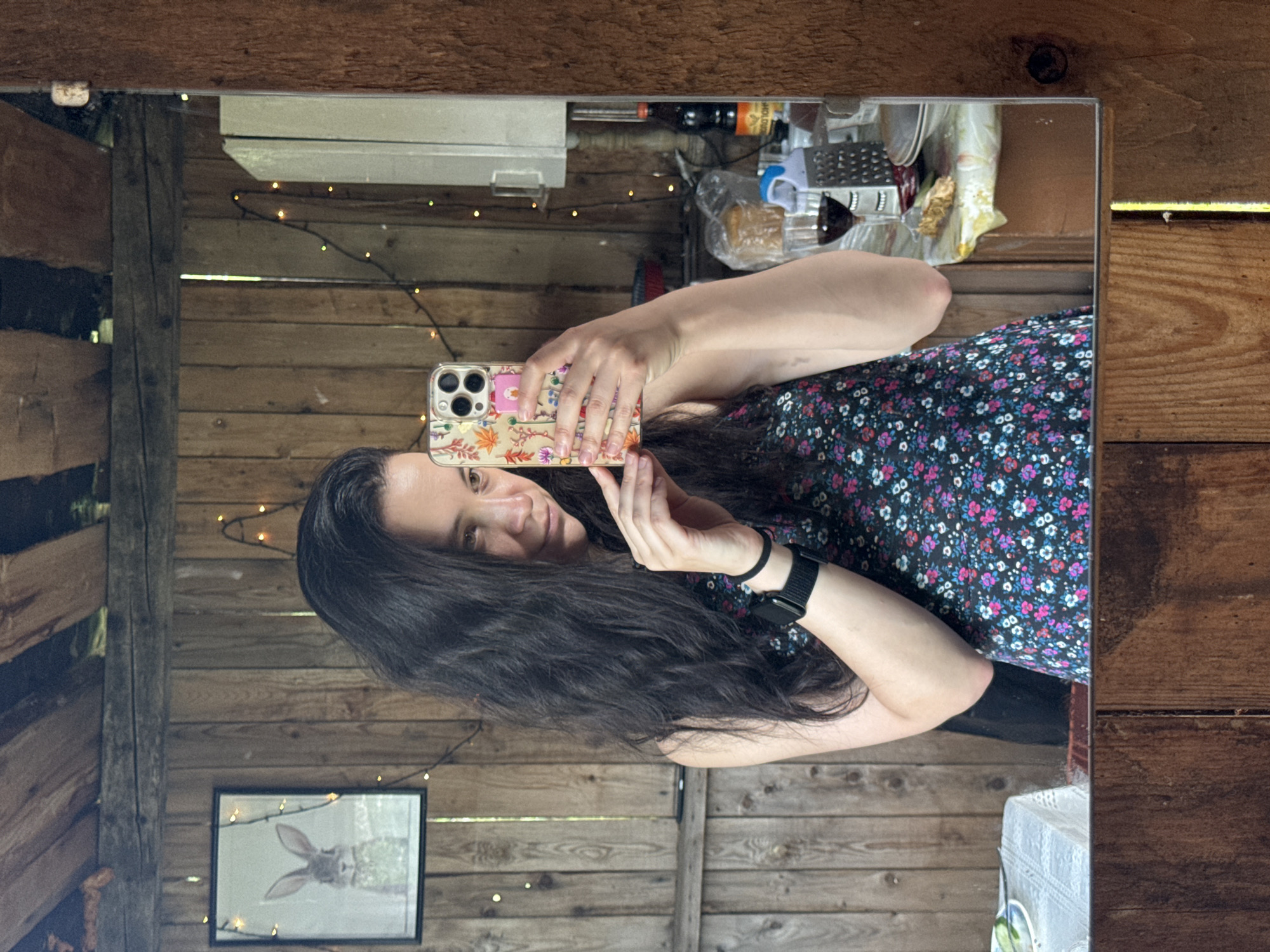 A woman with long, dark, wavy hair is taking a mirror selfie in what appears to be a rustic shed or outbuilding. She is wearing a black dress with a floral pattern of small pink, blue, and white flowers. On her left wrist, she wears a black smartwatch. In her right hand, she holds a smartphone with a colorful floral case.
The background is dominated by wooden plank walls and ceiling beams. String lights with small yellow bulbs are draped across the walls, adding a decorative element. To the left of the woman, a framed picture of a rabbit is hanging on the wall. To her right, a counter or table is visible in the mirror's reflection, with various items including a glass of red liquid, a grater, plates, and bottles. The floor is also made of wood. The lighting is dim, with the string lights providing some ambient illumination.
There is no specific information in the image to indicate the location is Zasechnyi, Russia.