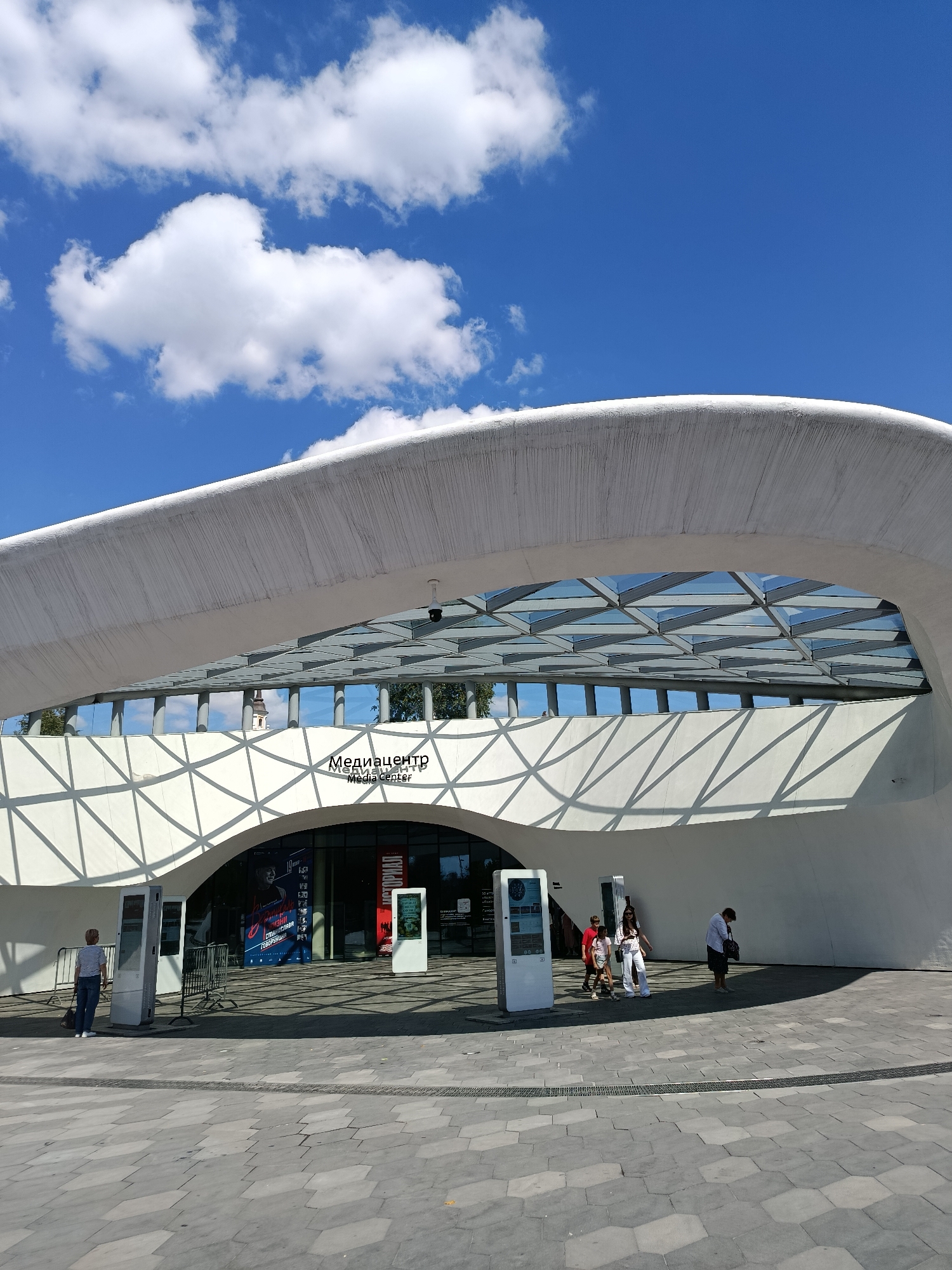 A wide shot captures the exterior of a modern building with a curved white roof and a glass ceiling, under a bright blue sky with scattered white clouds. The building features a large arched entrance with signage that reads "Медиацентр" and "Media Center" in Cyrillic and English. Several people are visible in front of the entrance. To the left, a woman in a striped shirt and jeans stands near a digital display. To the right, a group of people, including children, are walking. Another person, a woman in a white shirt and dark skirt, stands in the foreground on the right. The ground in front of the building is paved with hexagonal gray tiles. The scene appears to be a public space on a sunny day.