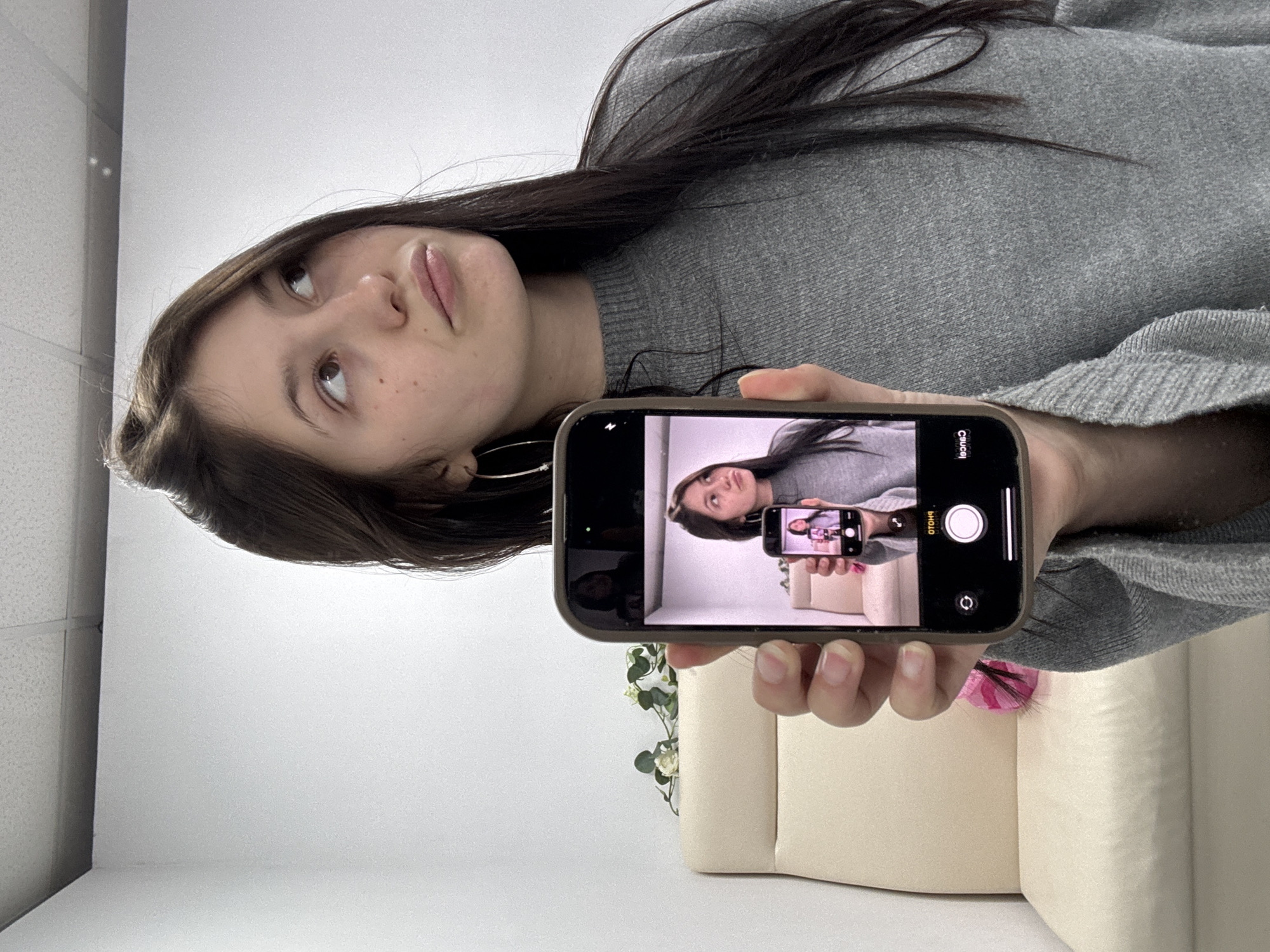 The image, originally rotated 90 degrees clockwise, depicts a young woman in a medium shot, holding a smartphone that displays a recursive image of herself. She has long, dark brown hair, fair skin, and is wearing a grey ribbed long-sleeved sweater. Her gaze is directed upwards and to her left, with a pensive or slightly contemplative expression. A silver hoop earring is visible on her left ear.
She holds a dark-cased smartphone in her right hand. The phone's screen shows its camera interface, with "PHOTO" selected and "VIDEO" as an alternative option, along with a shutter button and a camera switch icon. Crucially, the image on this phone's screen is a picture of the same woman, further holding a smaller phone that also shows an image of herself, creating a 'Droste effect'. On the screen of the phone depicted within the main phone, the word "Cancel" is visible on the bottom left and "PHOTO" on the bottom right, suggesting an active camera or preview mode.
The background is a simple, well-lit indoor space, featuring a plain white wall. In the upper left of the upright image, a white ceiling with a textured grid pattern is visible. Behind the woman's hand and the phone, parts of a cream or beige upholstered sofa or armchair can be seen. Artificial green foliage with small white flowers is partially visible near the sofa, along with a hint of a pink decorative item. The lighting is bright and even, characteristic of an indoor setting, possibly an office or commercial establishment in Фоки, Russia.