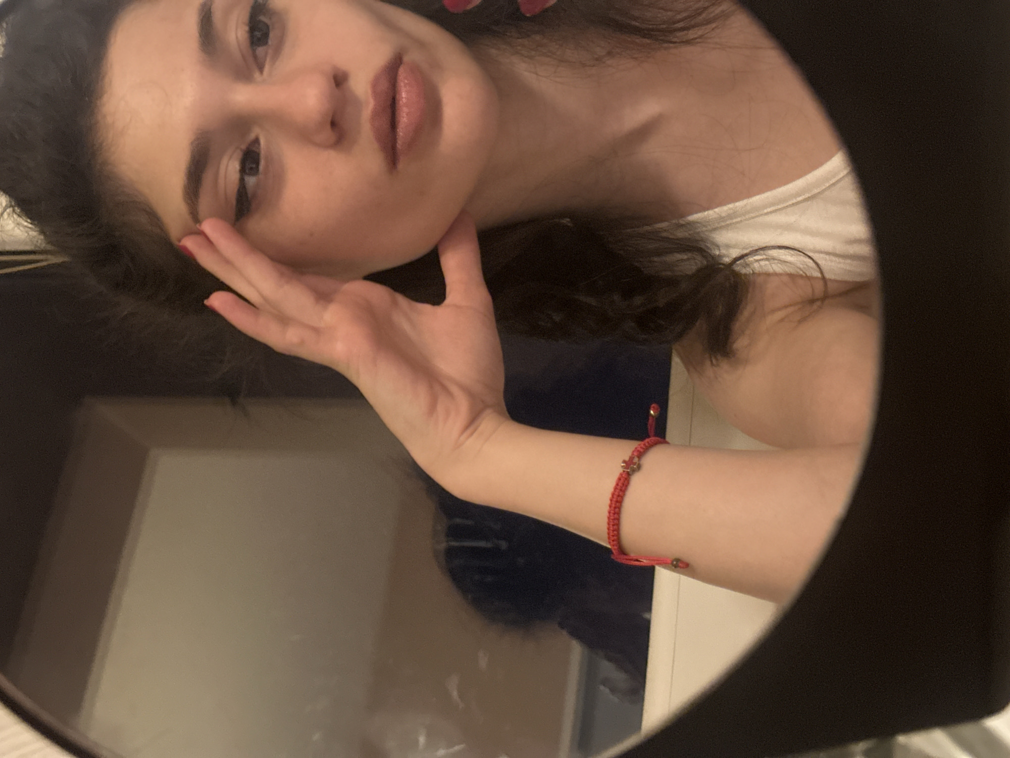 A young woman with dark curly hair and dark eyes is reflected in a round mirror. She is wearing a white tank top and a red string bracelet on her left wrist. Her left hand is raised to her face, and her fingers are touching her temple. She has winged eyeliner and a light pink lipstick on. The background of the reflection shows a blurred image of a room, with a dark object in the foreground that appears to be the edge of the mirror frame. The overall lighting is dim. The image is taken indoors, likely in a personal setting such as a bathroom or bedroom. There is no indication of the specific location within Sofia, Bulgaria.