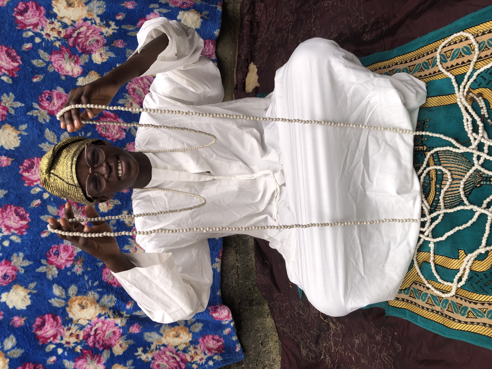 A young Black person wearing a white traditional outfit and a gold headwrap sits cross-legged on a patterned rug. The person is holding two long strands of white beads, one in each hand, extended upwards. The background is a blue fabric with a floral pattern of pink and yellow flowers. The person is wearing glasses and smiling at the camera. The scene appears to be outdoors, possibly in Ado, Nigeria, given the traditional attire and the style of the rug. The lighting suggests it is daytime.