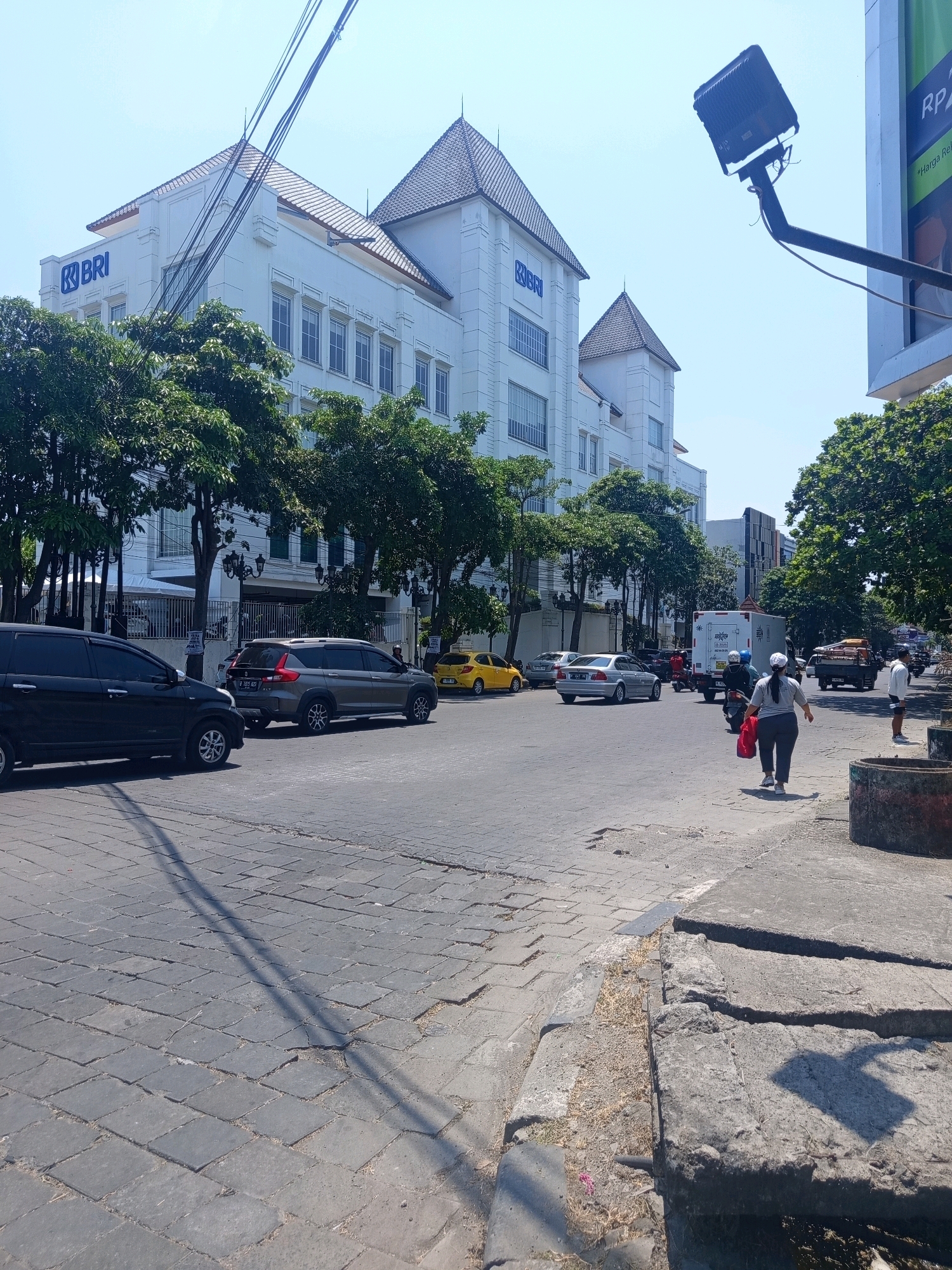 The image depicts a street scene in Semarang, Indonesia, under a clear blue sky. In the foreground, a paved street with cobblestones shows signs of wear and tear. To the left, a black SUV is parked, and a silver SUV is in motion further down the road. The middle ground features a grey SUV and a yellow car. Dominating the background is a large, white building with distinctive architectural features and the "BRI" logo prominently displayed, indicating it's a Bank Rakyat Indonesia branch. Trees line the sidewalk in front of the building. On the right side of the street, a woman in a grey shirt and dark pants, carrying a red bag, walks away from the camera. A white truck with a refrigerated cargo box is also visible on the road, along with other vehicles and pedestrians. A large, dark spotlight is mounted on a pole in the upper right of the image. The overall impression is of a typical urban environment on a sunny day.