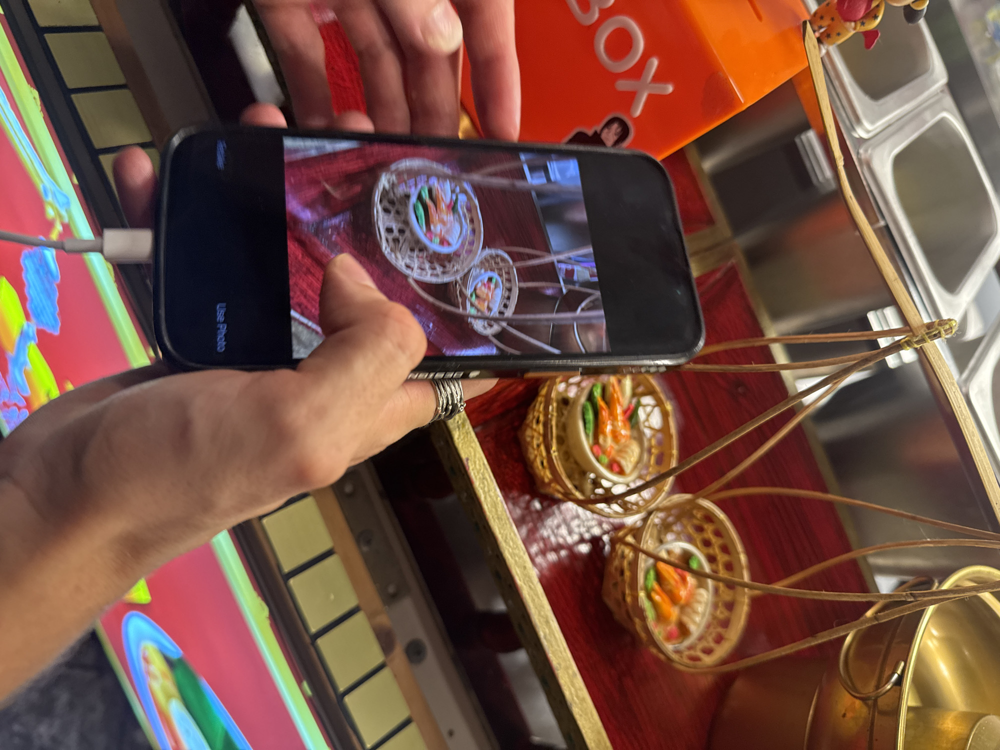 In Karon, Thailand, a person is holding a black smartphone, taking a picture of a miniature replica of a food vendor's stall. The stall features a wooden balance scale with two small, woven baskets hanging from it, each containing miniature food items. One basket has what appears to be shrimp and vegetables in a small bowl. In the background, there are stainless steel containers, a golden bucket, and an orange box with "BOX" written in white letters. The smartphone screen displays the live camera feed, showing the same miniature stall, with icons for "Retake" and "Use Photo" at the bottom. The scene is viewed from a slightly elevated angle, with the focus on the smartphone and the hands holding it.