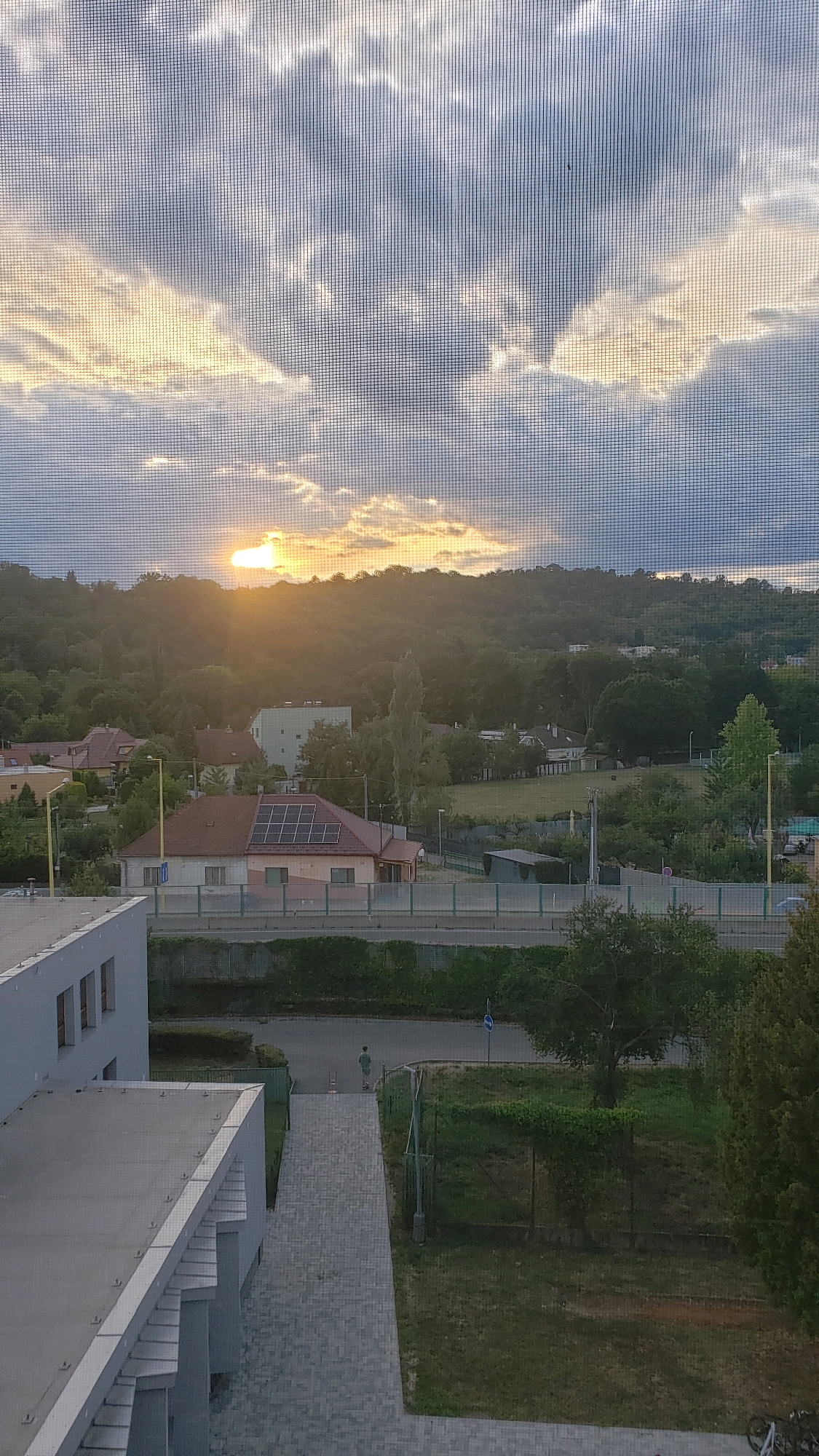 The image captures a view from a building overlooking a suburban landscape in Prešov, Slovakia, at sunset. In the foreground, a modern building's rooftop and facade are visible, constructed with light-colored materials and featuring geometric architectural elements. A paved pathway leads away from the building.
The mid-ground reveals a street lined with fences and trees. Across the street, a residential house with a red tile roof and solar panels is prominent, set against a backdrop of lush green hills. The hills are densely covered with trees, indicating a natural or park-like area surrounding the urban settlement. The sun is setting behind the hills, casting a warm glow and creating dramatic cloud formations in the sky.
In the distance, other buildings and more greenery are visible, suggesting a suburban or semi-rural environment within the city. A lone figure is walking on the pathway in the mid-ground, appearing as a small silhouette against the urban and natural elements. The overall atmosphere is serene, with the natural light of the sunset dominating the scene. The image is viewed through a window screen, which creates a subtle grid pattern across the entire frame.