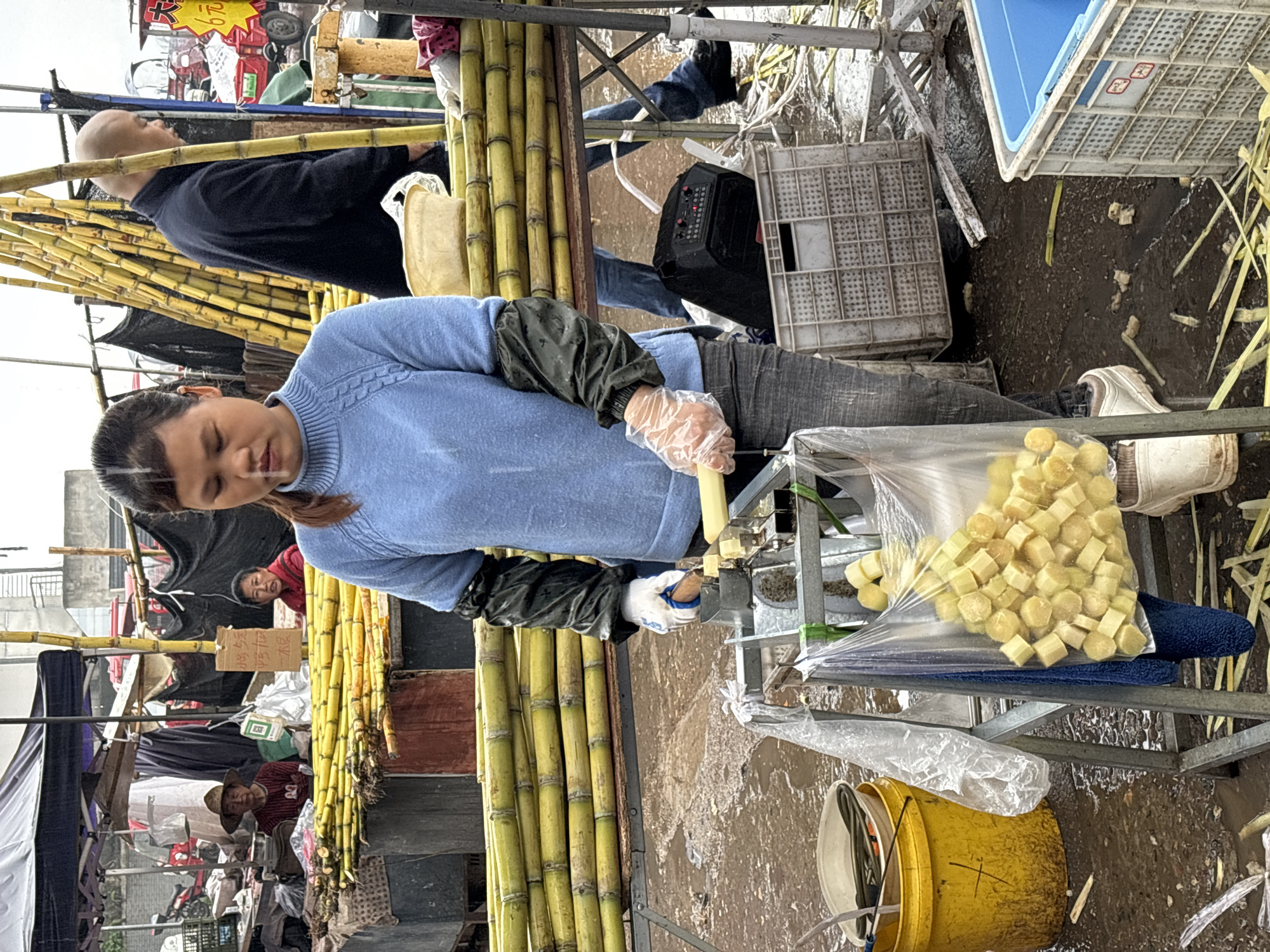 In Jiangjin, China, a woman with a ponytail and wearing a blue sweater and gloves operates a sugarcane cutting machine at an outdoor market. A stack of sugarcane stalks stands next to the machine. Cut sugarcane pieces are collected in a plastic bag. A bald man in a navy sweater stands nearby. The market has multiple stalls, and other people are visible in the background. The ground appears wet and muddy.