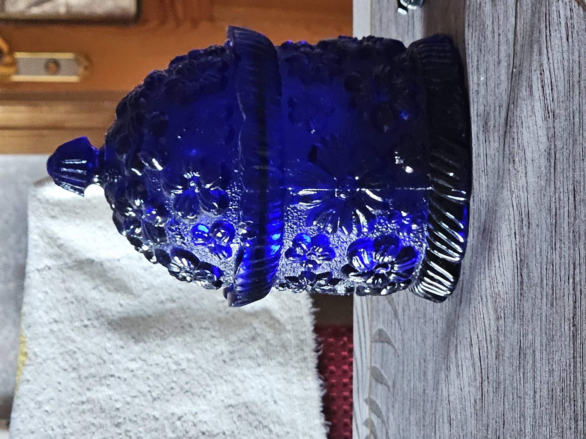 A cobalt blue glass candy dish with a lid is positioned on a weathered wooden table. The dish is elaborately decorated with raised floral patterns and has a fluted base. The lid features a finial also decorated with vertical grooves. In the background, a textured white fabric is visible, partially draped over a red surface. The environment appears to be indoors, with a wooden doorframe and wall visible to the right, suggesting a home or a decorative display setting. The lighting is somewhat dim, with light catching the facets of the glass.