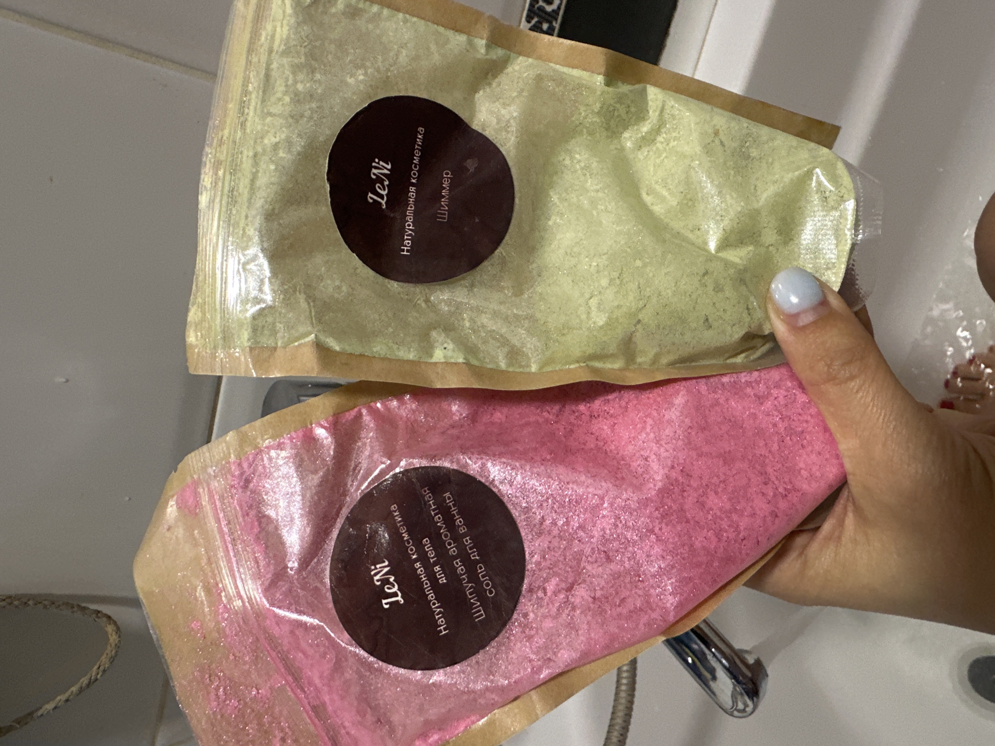 The image shows a person's hand holding two pouches of bath salts in a sink. The pouch on the left is pink and appears to contain pink bath salts. The pouch on the right is a light green color and contains green bath salts. Both pouches have circular labels with text in Russian. The text on the pink pouch reads "Le Ni, Натуральная косметика для тела, Шипучая ароматная соль для ванны". The text on the green pouch reads "Le Ni, Натуральная косметика, Шиммер". The sink is white, and a chrome faucet is visible with water running. The background appears to be a tiled wall. The location of the image is not discernible from the visual content and cannot be determined to be Novoaltaysk, Russia.
