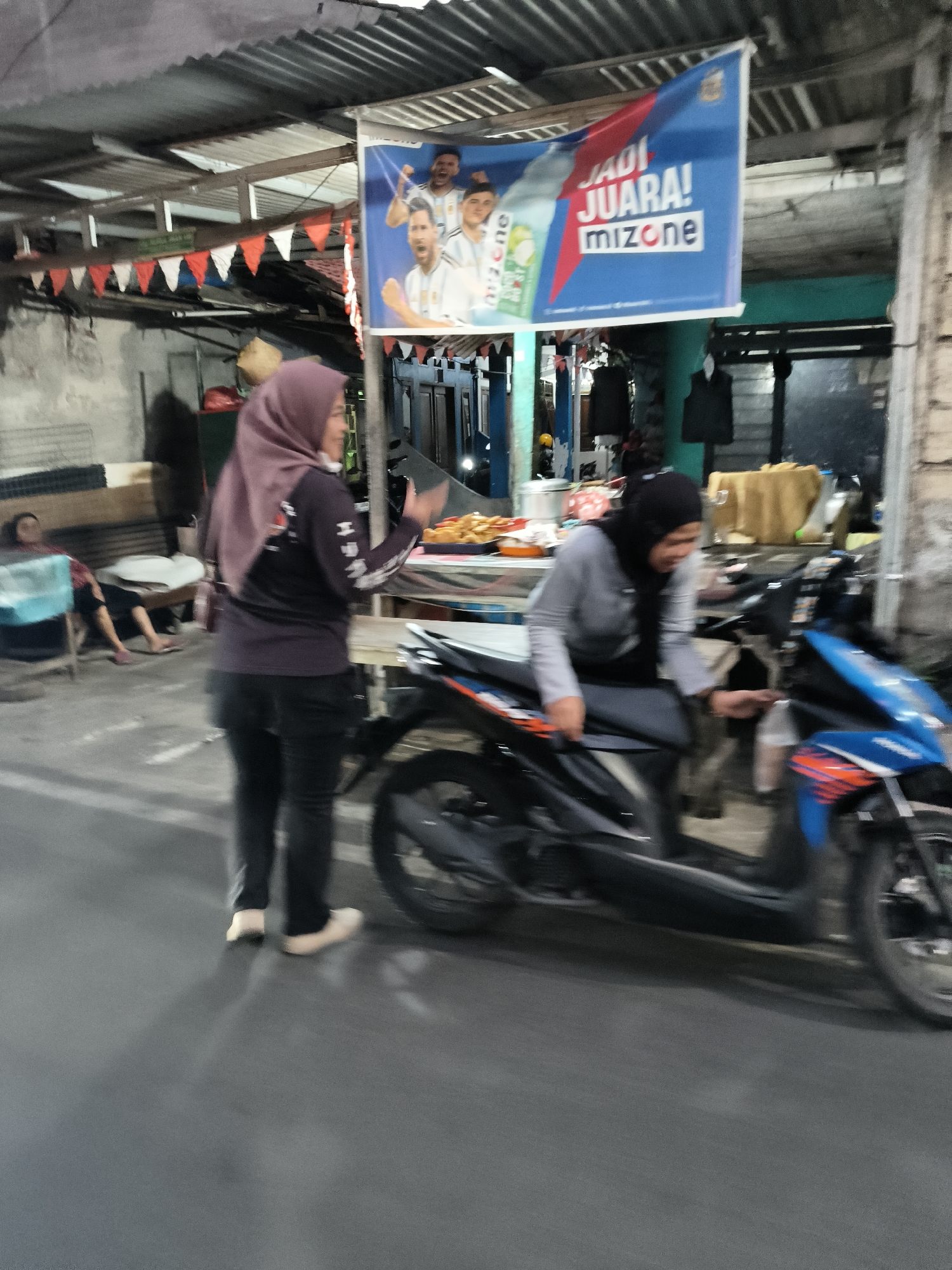 The image captures a street scene in South Tangerang, Indonesia, at what appears to be dusk or night. A blue banner hangs overhead, displaying text in Indonesian that reads "JADI JUARA! MIZONE" alongside images of soccer players, suggesting a promotional event or advertisement. In the foreground, two women are present. One woman, wearing a purple hijab and a dark long-sleeved shirt and black pants, stands facing a table with food items, holding out a hand as if to receive something. The other woman, wearing a black hijab and a grey long-sleeved shirt and grey pants, is seated on a scooter. She is reaching towards a small plastic bag. A motorcycle is parked in front of her. In the background, other individuals are seated, and there are structures that suggest a market or roadside stalls. The environment appears to be outdoors, with a corrugated metal roof visible above.
