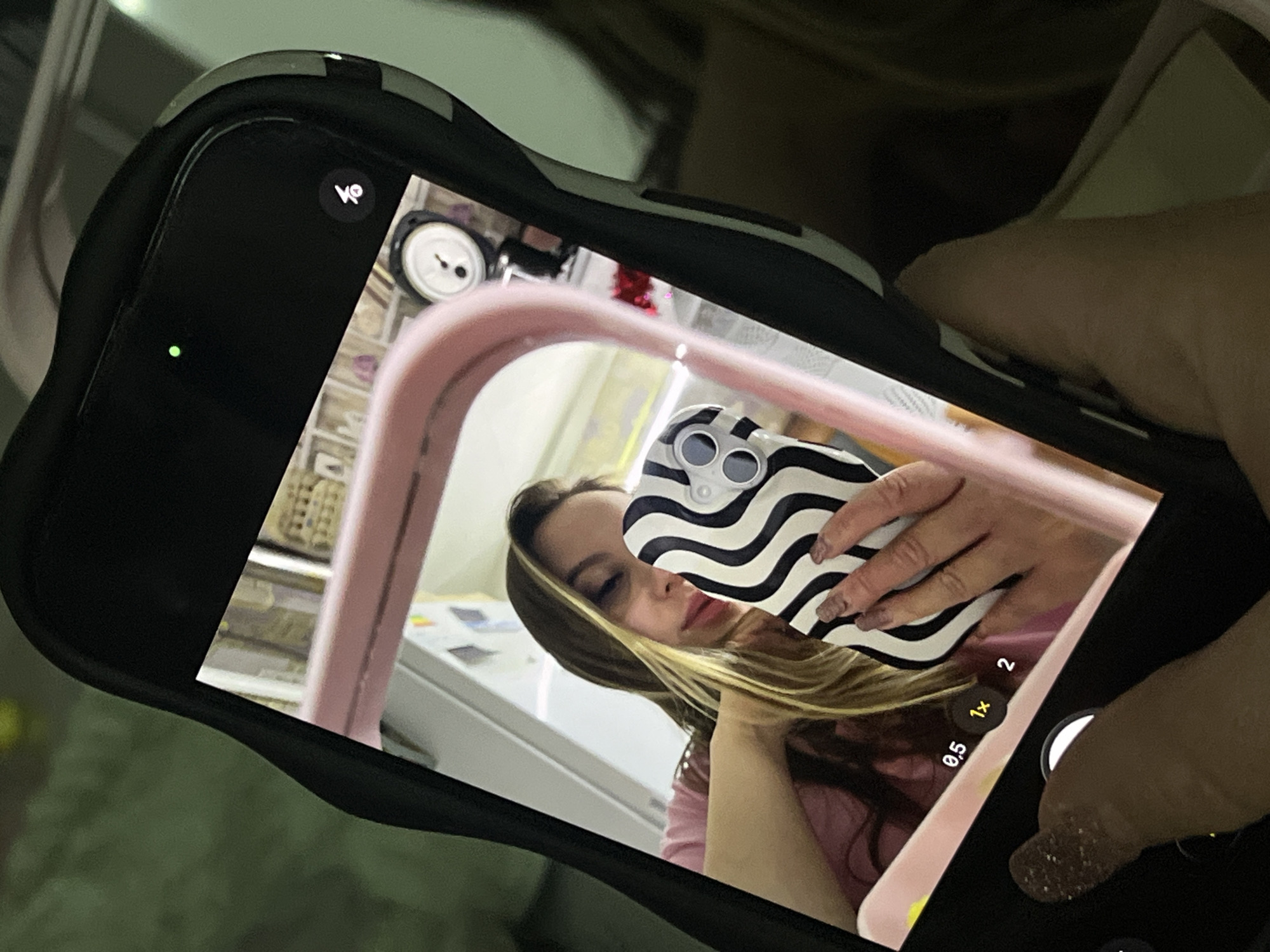 A person is holding a phone and taking a picture of a reflection in a mirror. The reflection includes the person holding the phone, a black and white wavy patterned phone case, and a pink structure in the background. There are also items on shelves in the background, out of focus. The location in the city of Port-Katon, Russia is not evident from this image.