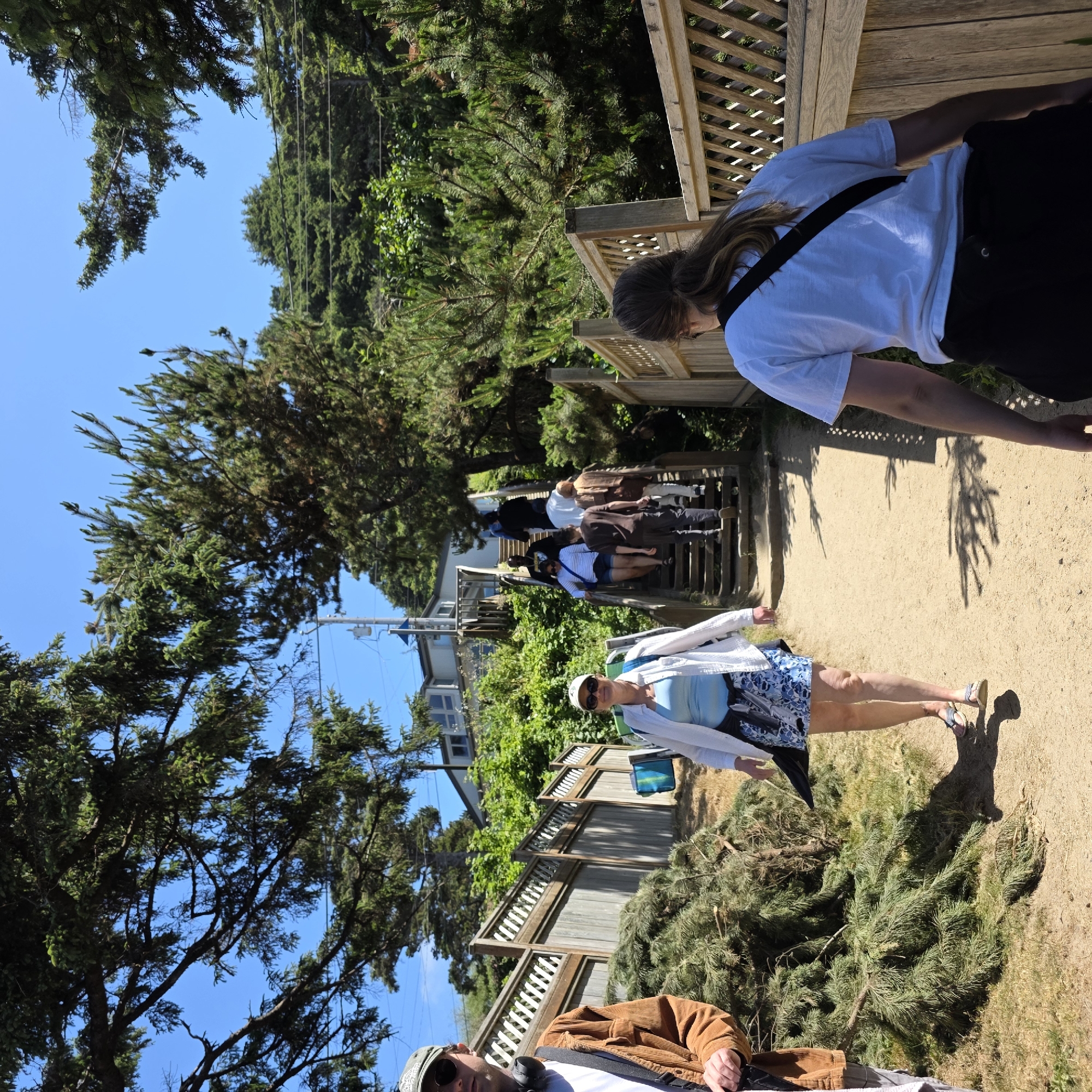 A group of people are ascending a wooden staircase and a dirt path in Cannon Beach, Oregon, USA. In the foreground, a woman with sunglasses and a white shirt over a blue tank top, wearing a blue and white patterned skirt and sandals, walks on a dirt path. To her left, a pile of pine branches is situated next to a wooden fence. A man wearing a brown corduroy jacket, sunglasses, and a cap is partially visible to the far left. Further up the path, a woman in a white shirt and a patterned skirt walks on the dirt path. Beyond her, several people are walking up the wooden stairs. The staircase leads to a house with a light-colored exterior, nestled among lush green trees and foliage. The sky is clear and blue. The wooden fence runs along the left side of the path. Another wooden fence is visible on the right side of the path, with a person in a white t-shirt facing away from the camera walking alongside it.