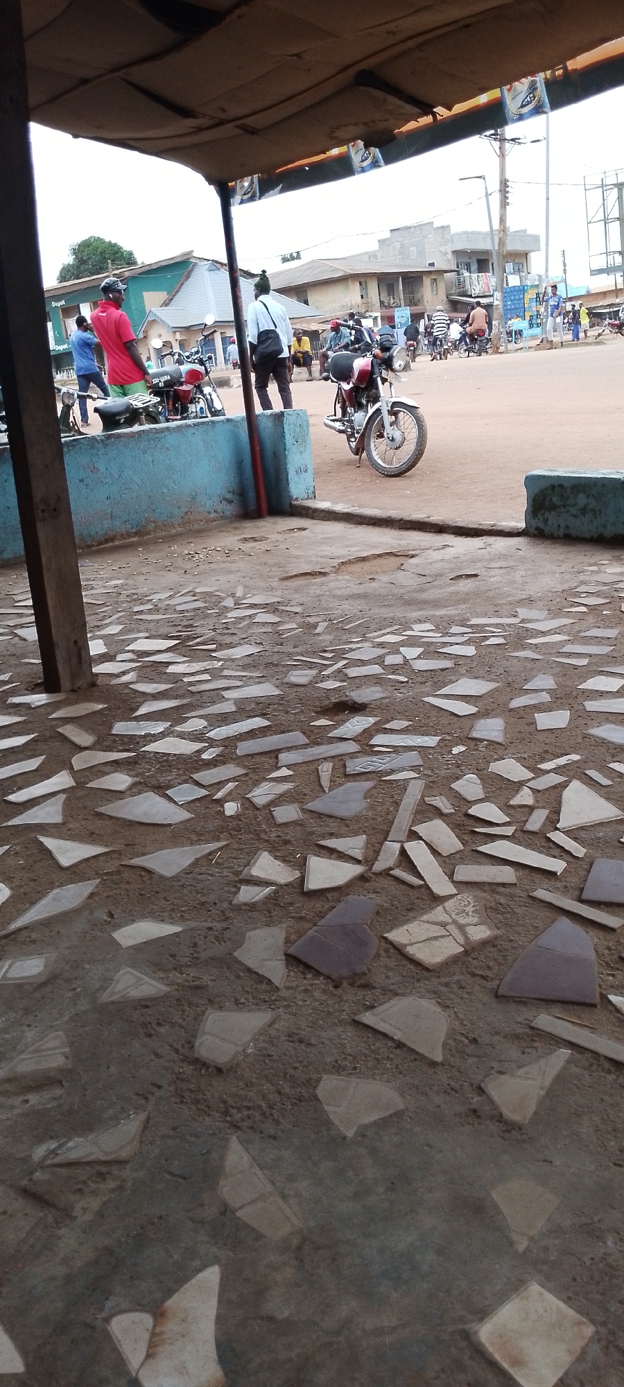 The image captures a scene in Gboko, Nigeria, likely a roadside area with a small shelter. In the foreground, a ground surface covered with broken pieces of tile is visible. Behind this, a blue concrete barrier runs horizontally. Several motorcycles are parked along the barrier.
In the middle ground, a person in a red shirt and black cap is standing, facing away from the camera. To their right, another person in a white shirt and dark pants, carrying a bag, stands looking towards the right. Further to the right, a red and silver motorcycle is parked.
In the background, other people are visible near parked motorcycles and in front of buildings. The buildings appear to be of simple construction, consistent with an urban or semi-urban environment in Nigeria. The overall atmosphere suggests a public gathering or waiting area, possibly near a transportation hub or market. The sky is overcast.