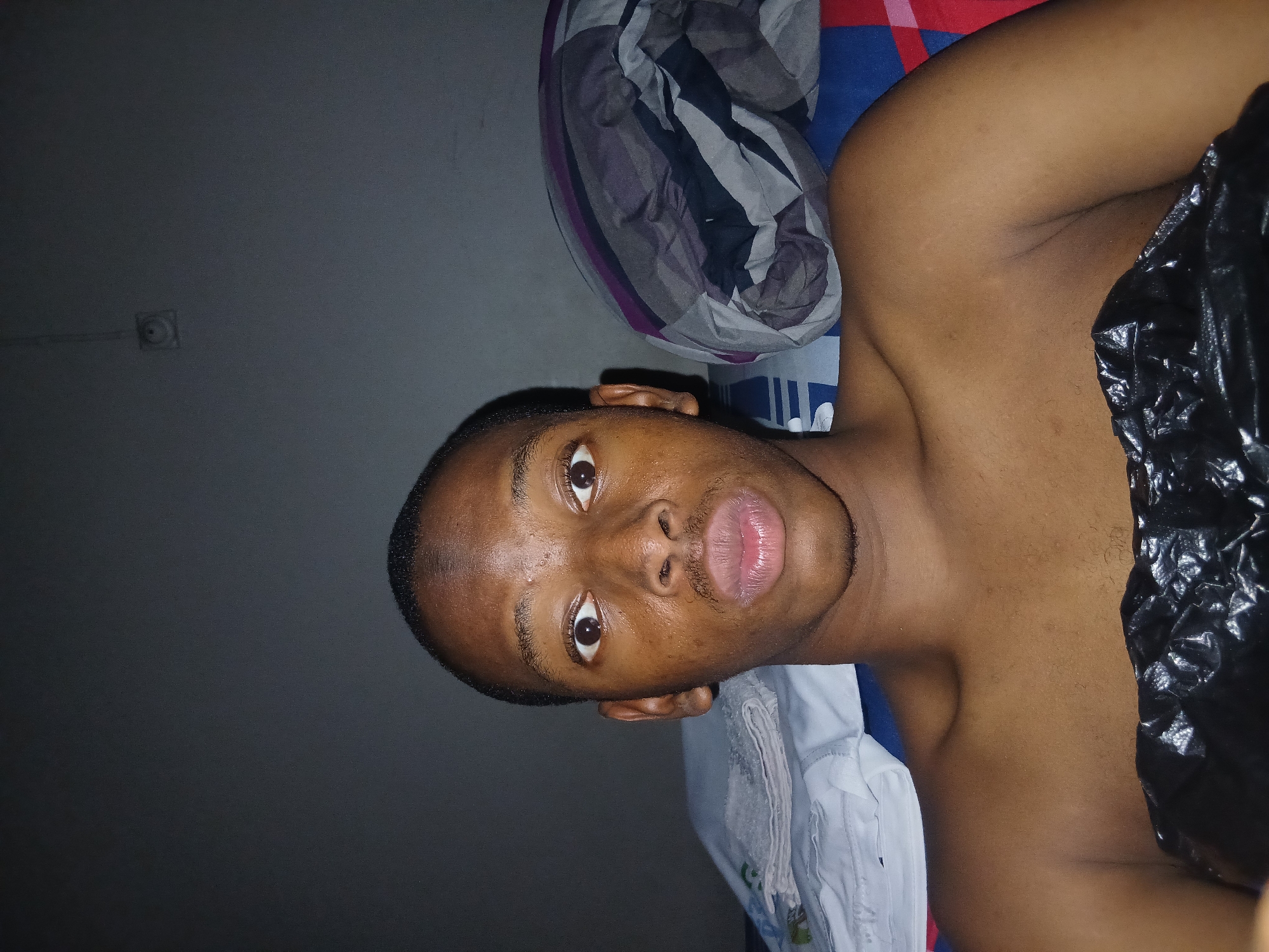 The image is a close-up, low-angle selfie of a young Black male with dark skin and short hair. He is lying down, and the shot focuses on his face and upper chest. His eyes are wide open, and his lips are pursed. He appears to be indoors, in what looks like a bedroom. Behind him, on the left, is a stack of folded white towels or cloths on a bed. To his right, a pillow with a patterned cover is visible. The background wall is a dark, neutral color, and a light fixture or socket is visible on the wall in the upper right quadrant. A black plastic material covers the lower part of his torso. The scene is dimly lit, suggesting it was taken at night or in a dark room. The presence of a pillow and bedding indicates a domestic setting, consistent with a residential area in Lagos, Nigeria.