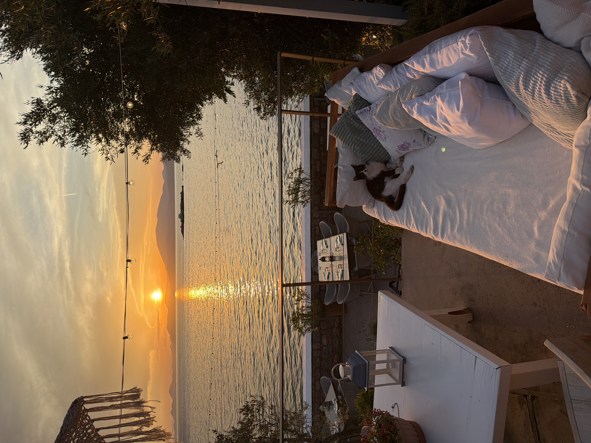 Sunset at Hydra, Greece. A view of the coastline with the sun reflected in the water. There is a daybed with white linens, pillows, and a sleeping cat. A small white table with a lantern and a dinner table with place settings. Trees and foliage are in the foreground.