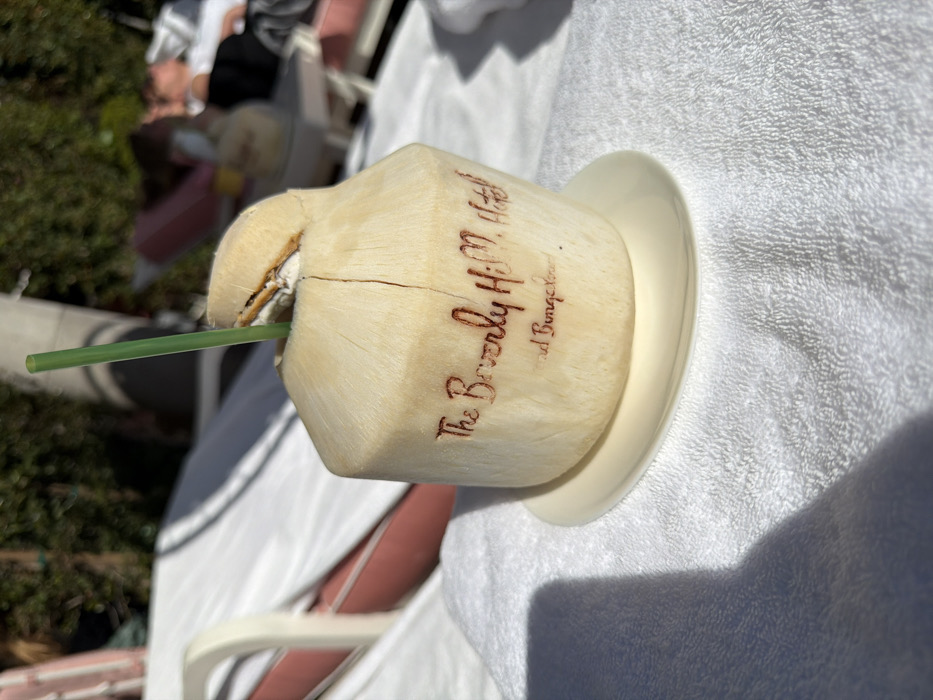 A fresh coconut drink, likely from the Beverly Hills Hotel, sits on a saucer atop a white towel. The coconut is husk-on, carved into a squared shape, with the hotel name burned into its side. A green straw protrudes from the top. The scene appears to be outdoors, near a lounge chair, suggesting a sunny, relaxed setting.