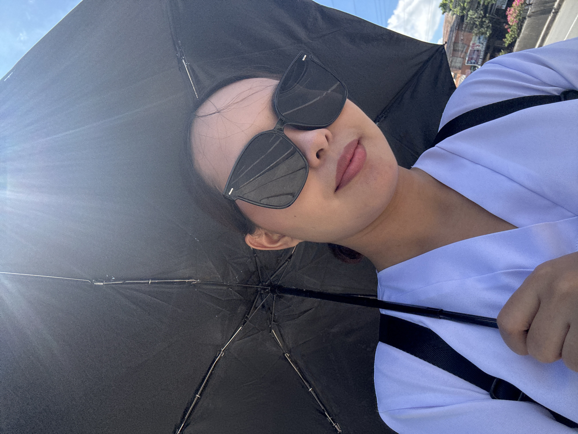 A medium-shot photograph depicts a person holding a black umbrella, presumably for shade, in Davao City, Philippines. The person, wearing sunglasses, is positioned in the foreground, and appears to be wearing a white shirt with black straps. The handle of the umbrella is visible in the person's grip. The background shows a glimpse of the city, including buildings and foliage, under a sky with partial cloud cover. A strong light source, possibly the sun, is situated to the left of the image.