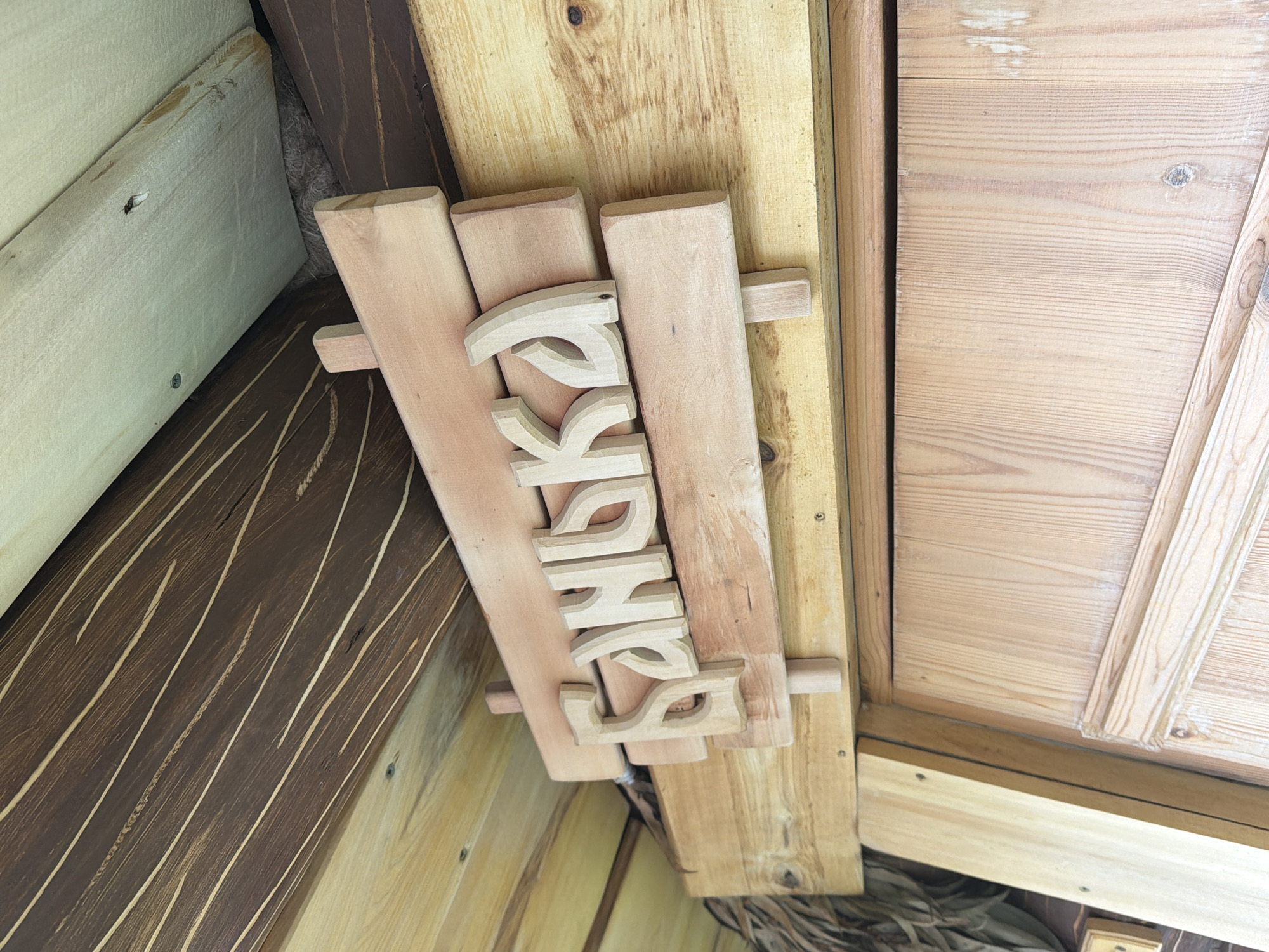 The image displays a close-up view of a wooden sign with carved letters that spell out "SHUSHA". The sign is attached to a light-colored wooden plank, which is itself mounted on a darker wooden surface. The carving style is rustic, with rounded edges on the letters. The sign appears to be outdoors, as indicated by the natural wood grain and the presence of some dried vegetation or straw visible beneath the sign. The background consists of wooden planks with varying wood grain patterns and colors, suggesting a natural or rustic construction. There is no indication of time of day or weather conditions, and no people are visible in the image. The context provided places the location as Nizhnyaya Shilovka, Russia.