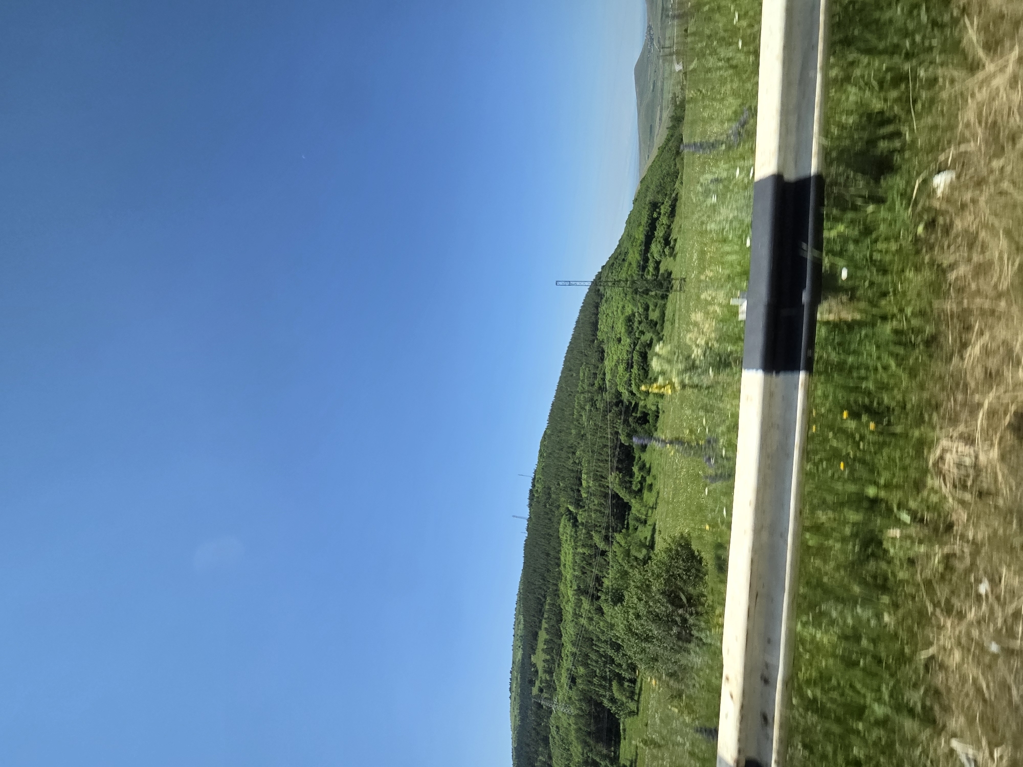 The image shows a hilly, green landscape under a clear blue sky. A white and black guardrail is in the foreground, with blurred greenery and dry grass beneath it. In the mid-ground, a lush green hillside is covered with trees and grass, with several tall, slender transmission towers visible on its slope. In the distance, rolling hills fade into the horizon. There are no people or animals visible in the image. The location is the city of Krashen, Armenia.