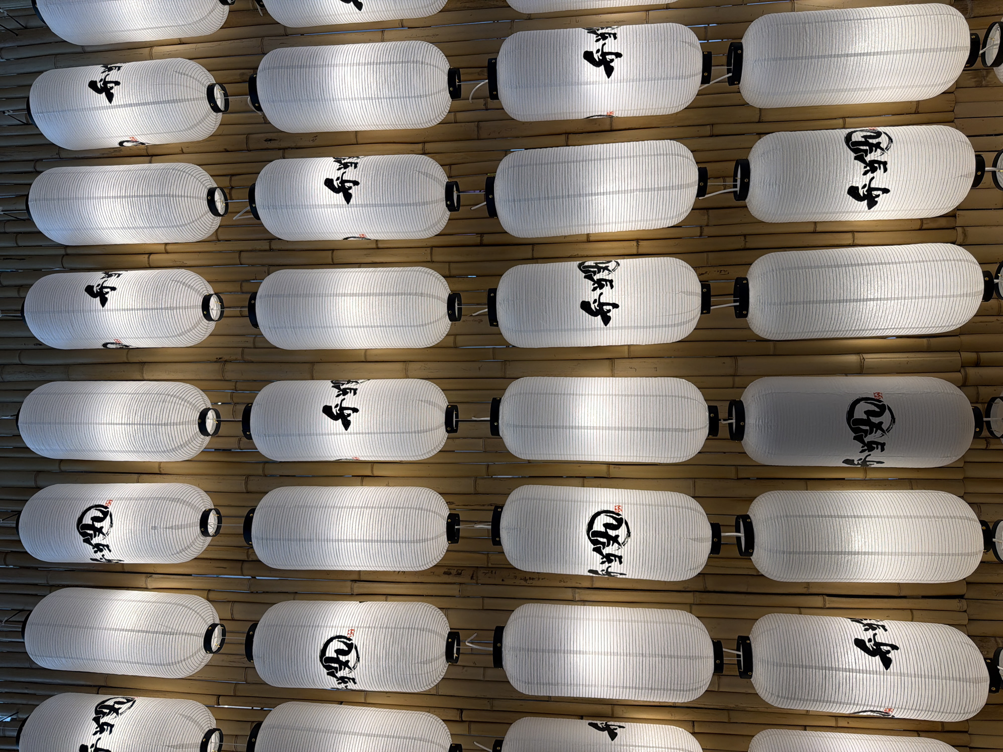 The image displays a vertical arrangement of white cylindrical paper lanterns, each adorned with black Japanese calligraphy. The lanterns are suspended against a backdrop of vertical bamboo stalks. The calligraphy on the lanterns appears to be consistent across most of them, with a circular emblem above a larger character. The lighting suggests an indoor setting, possibly a restaurant or decorative space. There are no people visible in the image. The scene depicts a decorative installation of Japanese lanterns.