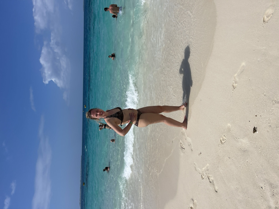 A tropical beach scene on a sunny day in the city of Zanzibar, Tanzania. A young adult female stands barefoot on light-colored sand in the foreground, facing slightly left with her head turned towards the camera, smiling. She wears a two-piece bikini with a brown and black animal print, and her hands are positioned on her hips. Her blonde hair appears wet. Gentle waves break near her feet, and visible footprints mark the sand. To her right, a distinct human shadow is cast on the wet sand. In the turquoise ocean water behind her, approximately seven individuals are partially submerged, with only their heads and shoulders visible. The water extends to a clear horizon under a bright blue sky with scattered white clouds. A distant landmass is visible on the left horizon, and a yellow buoy floats in the water in the mid-distance.