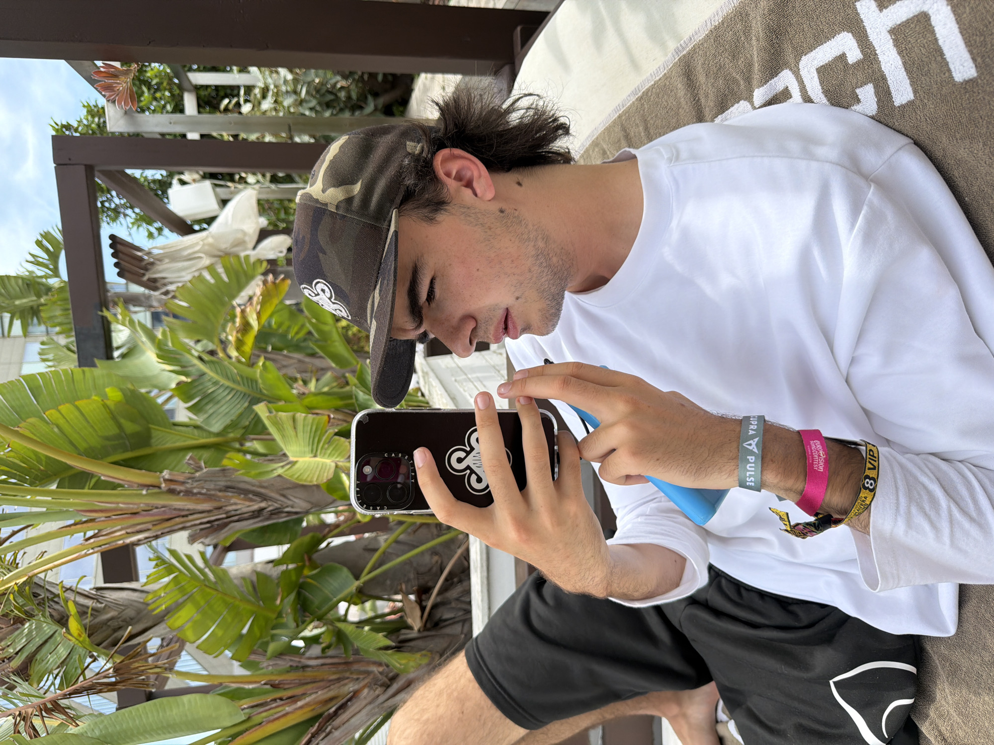 The image depicts a young male, appearing to be in his late teens or early twenties, relaxing outdoors at what appears to be an upscale hotel or resort in Barcelona, Spain. He is lying on a brown towel, which features a large white letter 'H' as part of its branding, on a sun lounger or outdoor bed.
The male has dark hair styled in a mullet, with visible stubble on his face. He wears a camouflage baseball cap with a white stylized logo on the front. His attire includes a white long-sleeved t-shirt and black shorts. On his right wrist, he wears multiple bands: a grey-green band with "SUPRA PULSE", a pink band with "EUROVISION SONG CONTEST MALMÖ", and a yellow-black band reading "AMNESIA IBIZA 8 VIP".
He is intently focused on a black smartphone, possibly an iPhone, held in his left hand. The phone is encased in a clear Casetify brand case, which displays a white stylized clover-like graphic on the back and accommodates a triple camera system. His right hand holds a slender blue cylindrical object, possibly a vape pen or stylus, which is positioned towards the phone screen.
The background features lush tropical foliage, predominantly large-leafed plants resembling Bird of Paradise species, suggesting a warm climate. Behind the vegetation, a modern, multi-story building with glass facades is visible. The scene is set under a bright blue sky with scattered white clouds. A dark brown wooden pergola structure is visible above and behind the male, with some white fabric elements hanging from it, possibly decorative or part of a shading system. The overall environment conveys a leisurely, sunlit outdoor setting.